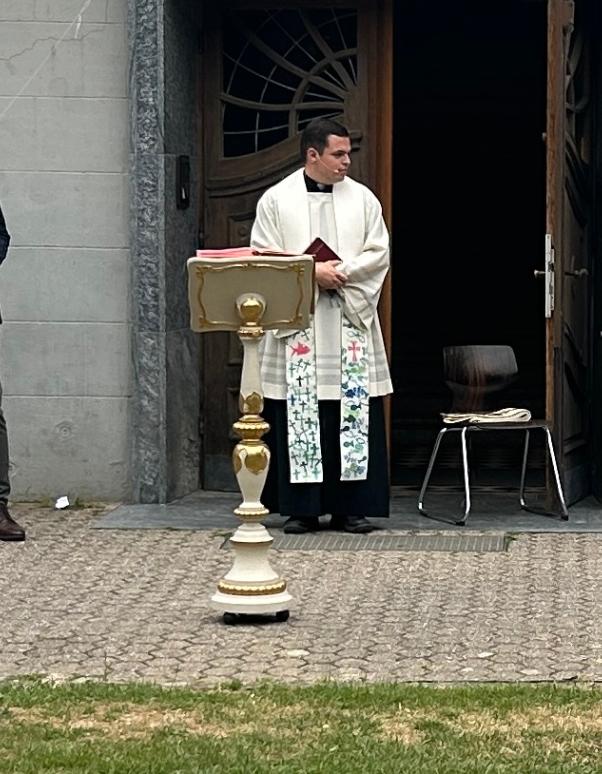 Priester mit bunter Stola in der Mitte, links ein Holzesel, daneben ein Mann, rechts zwei Frauen