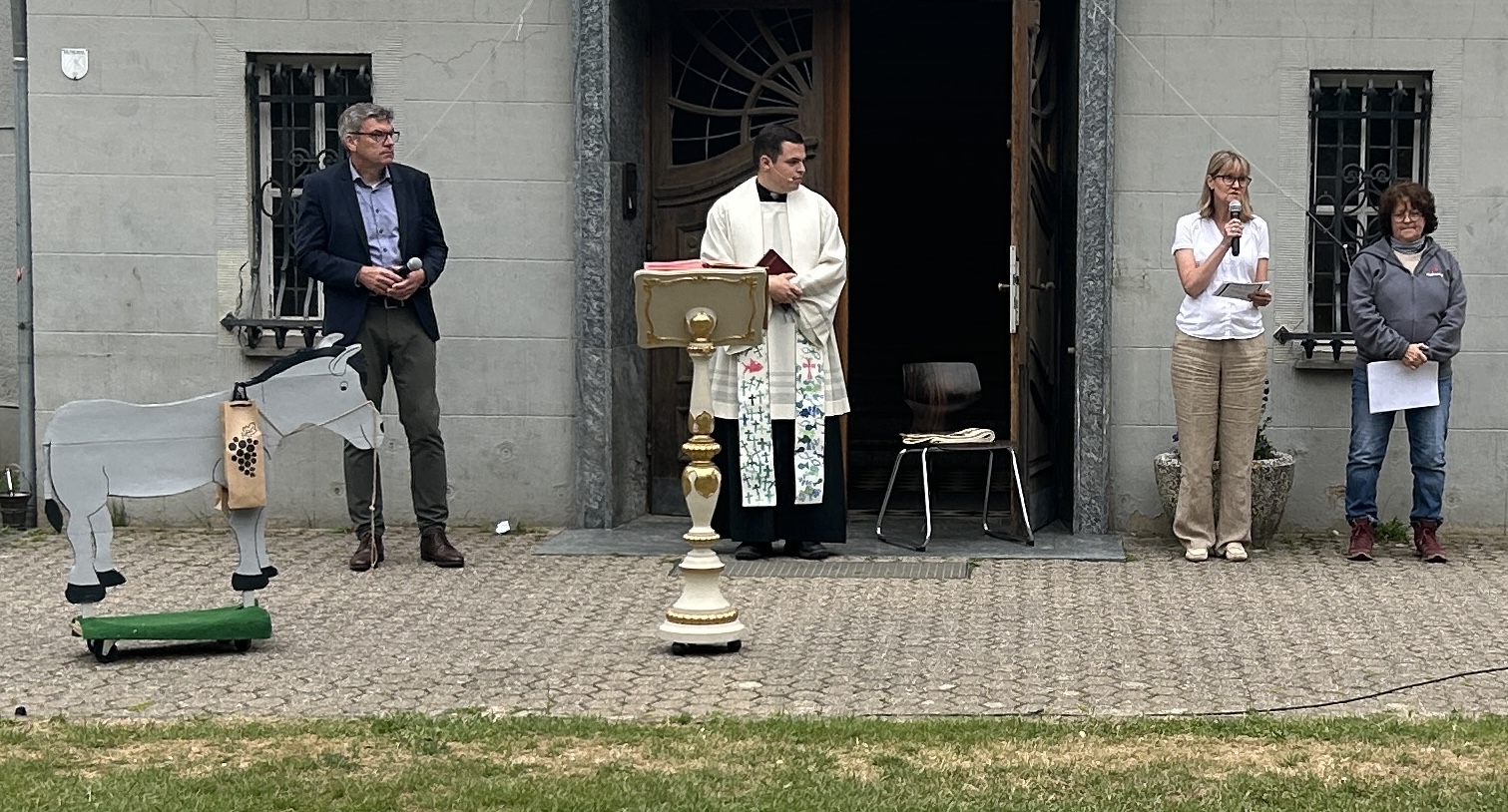 Priester mit bunter Stola in der Mitte, links ein Holzesel, daneben ein Mann, rechts zwei Frauen