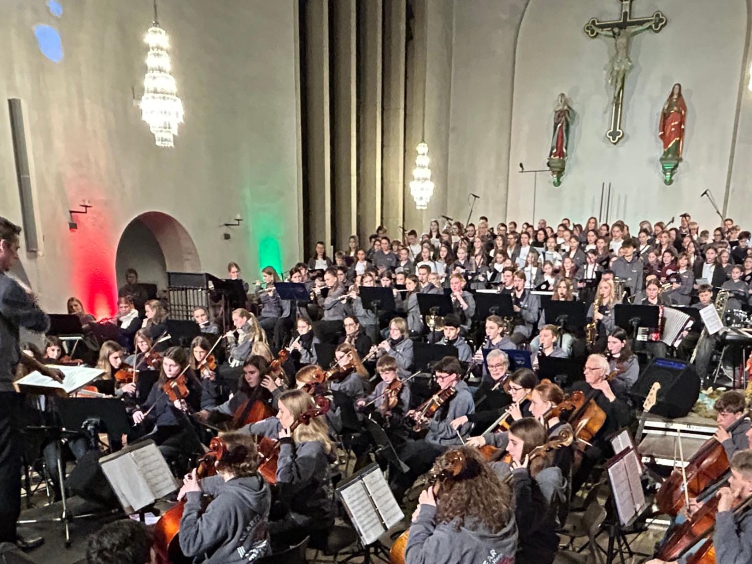 Orchester aus Kindern, Jugendlichen, Erwachsenen in einer Kirche, dahinter Kinder, Jugendliche Erwachsene im Chor, vorne Dirigent, farbige Lichter