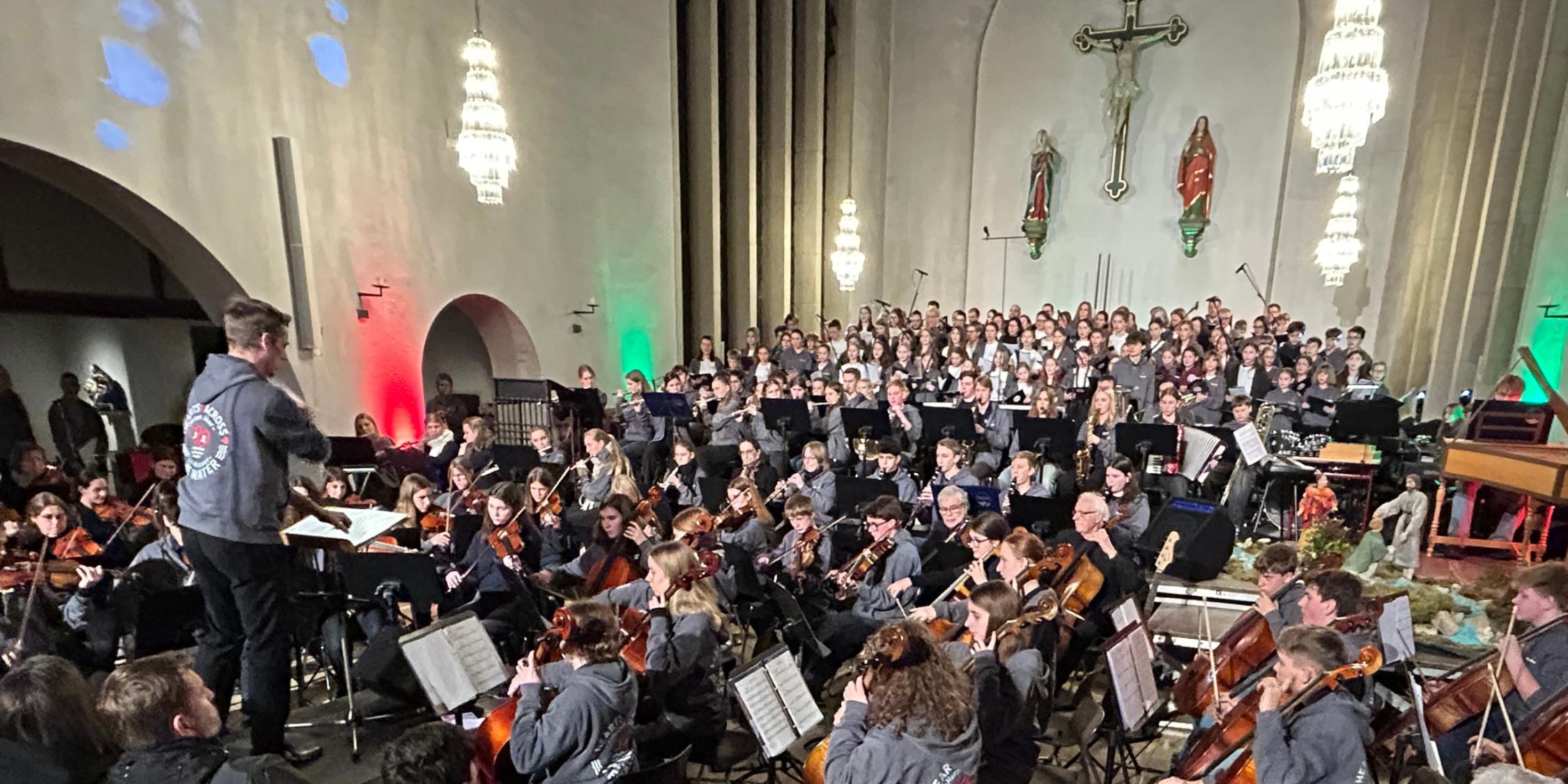 Orchester aus Kindern, Jugendlichen, Erwachsenen in einer Kirche, dahinter Kinder, Jugendliche Erwachsene im Chor, vorne Dirigent, farbige Lichter
