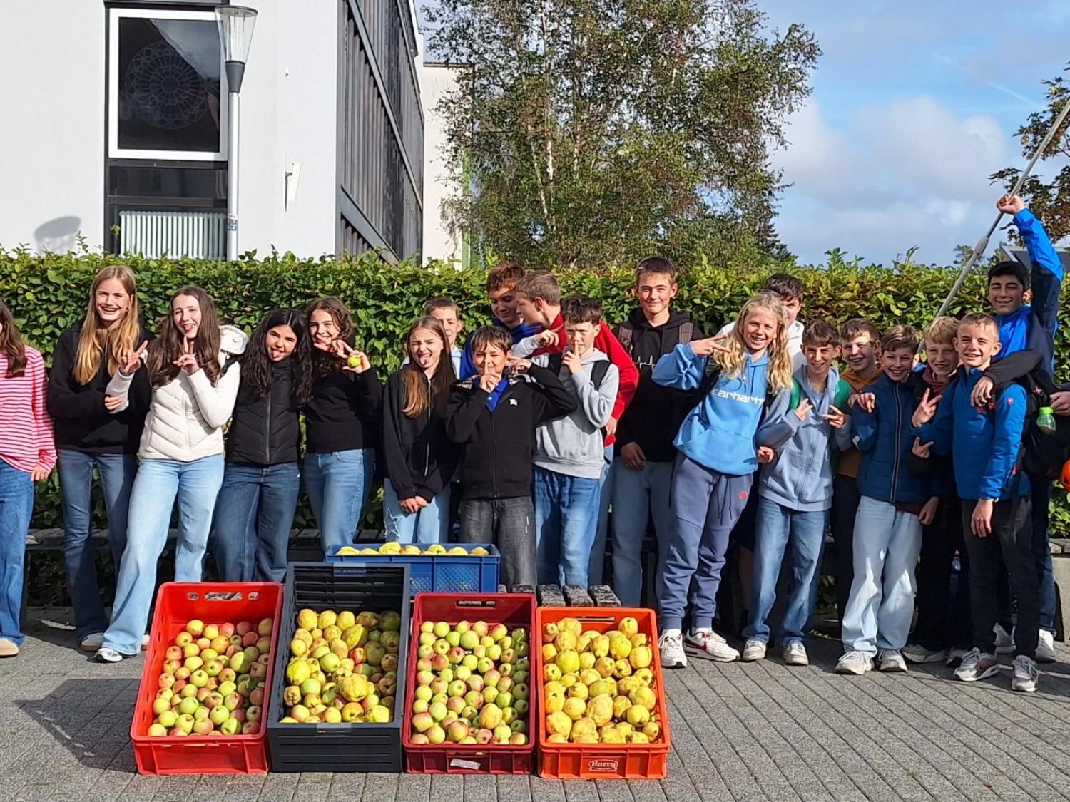 Schülerinnen und Schüler nebeneinander vor dem Schulgebäude, Obstpflücker in der Hand, davor vier Kästen mit Äpfeln und Quitten