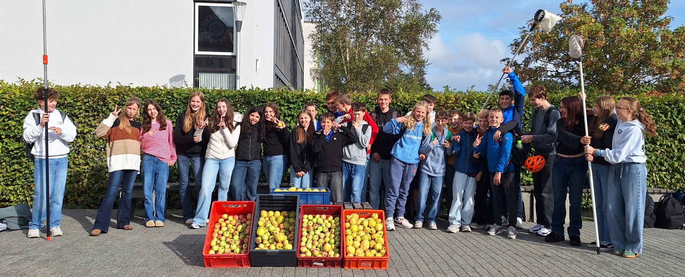 Schülerinnen und Schüler nebeneinander vor dem Schulgebäude, Obstpflücker in der Hand, davor vier Kästen mit Äpfeln und Quitten