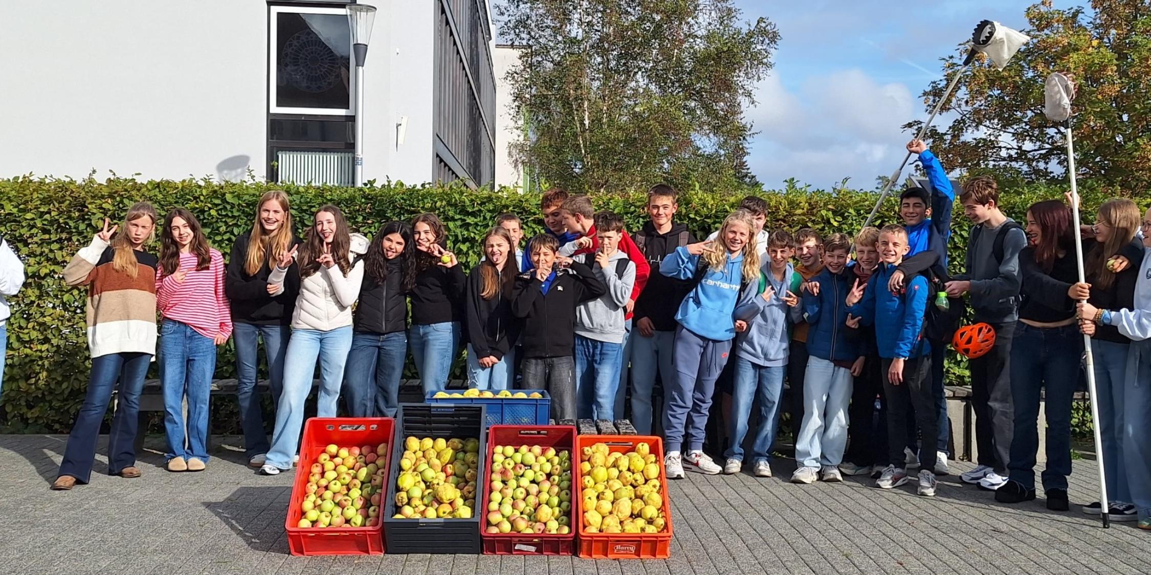 Schülerinnen und Schüler nebeneinander vor dem Schulgebäude, Obstpflücker in der Hand, davor vier Kästen mit Äpfeln und Quitten
