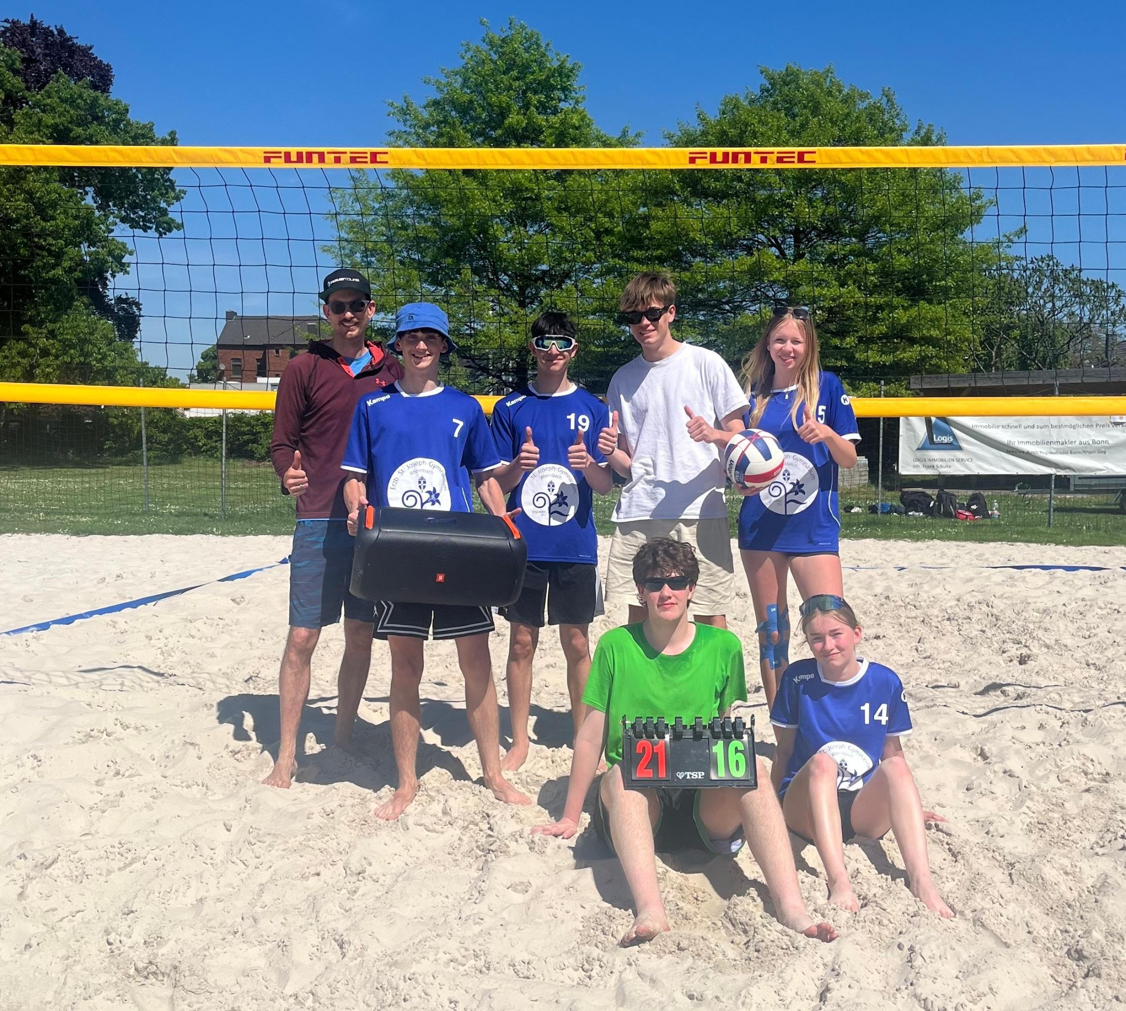 acht Volleyballspieler:innen stehen und sitzen vor einem Netz im Sand