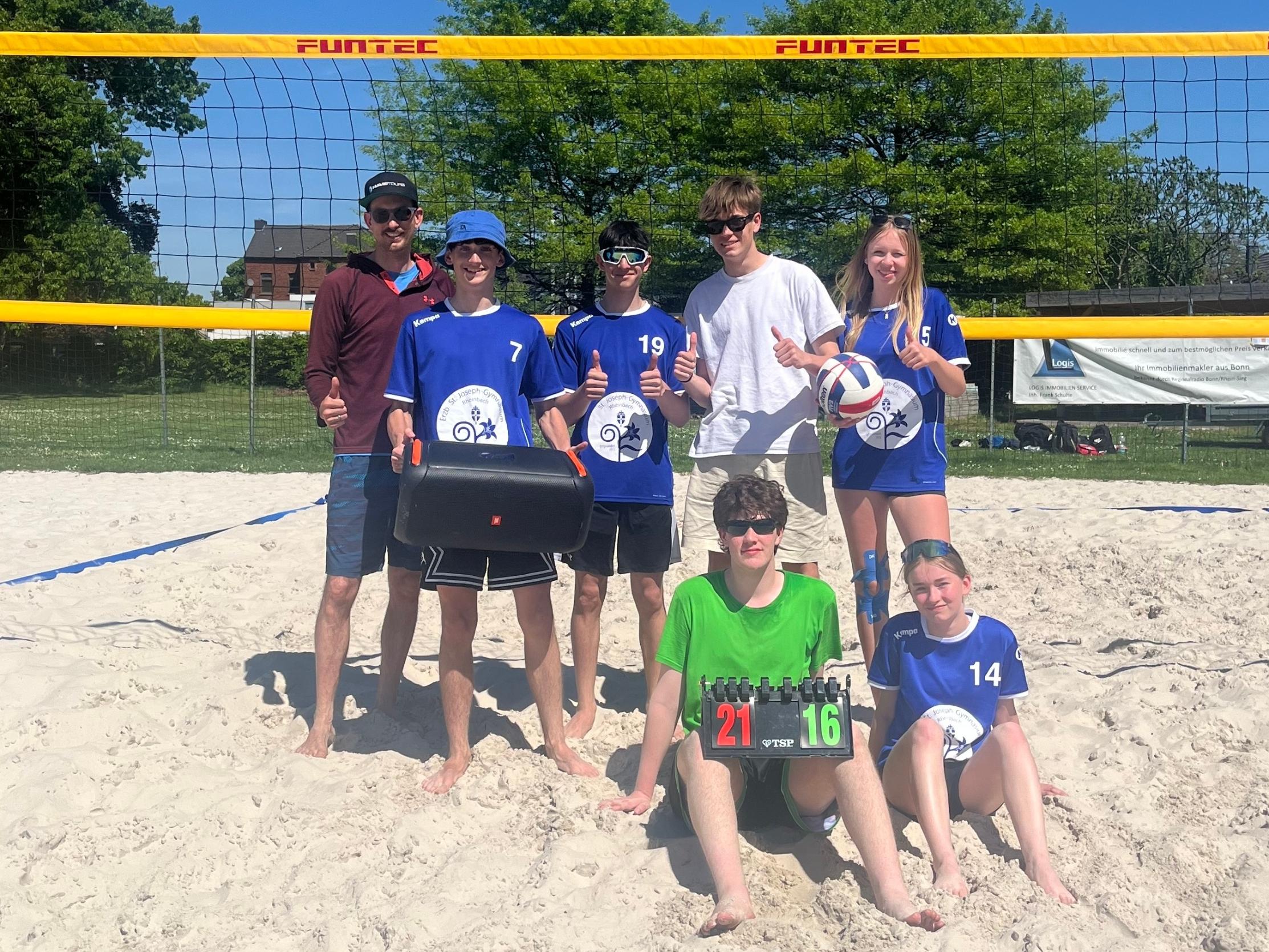 acht Volleyballspieler:innen stehen und sitzen vor einem Netz im Sand