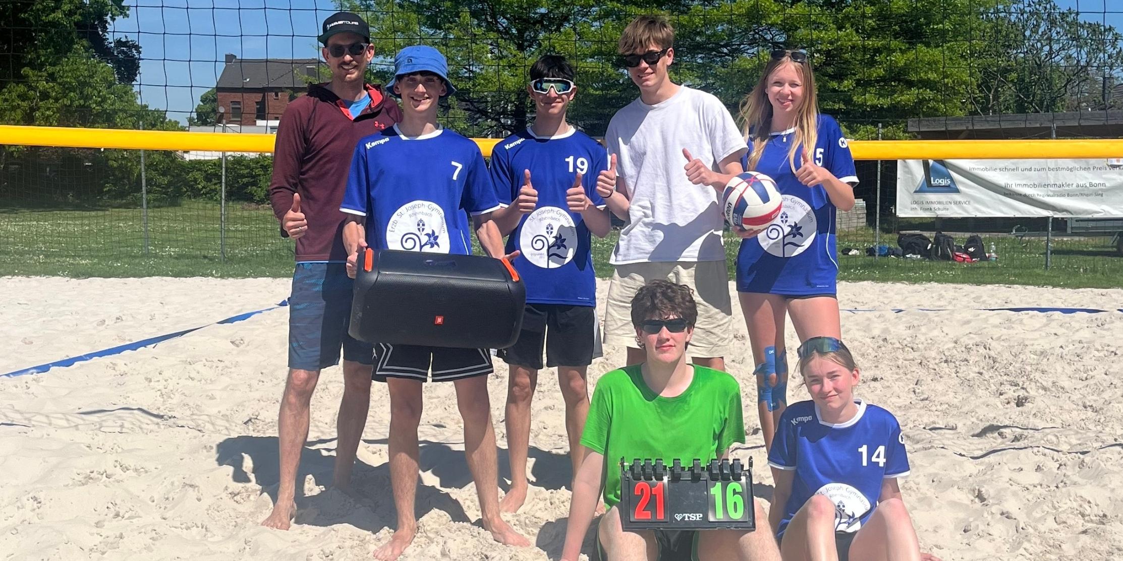 acht Volleyballspieler:innen stehen und sitzen vor einem Netz im Sand