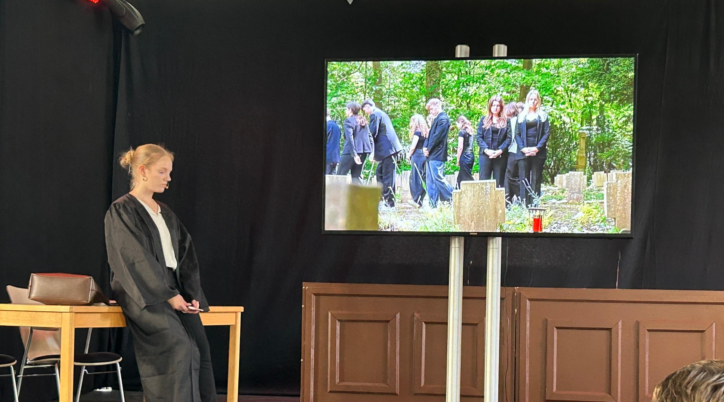 Monitor, darauf Beerdingunsszene, daneben die gespielte Rechtsanwältin