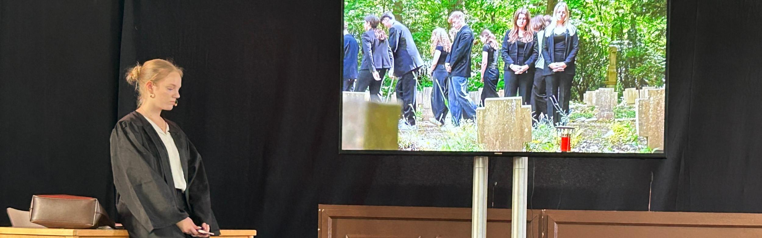 Monitor, darauf Beerdingunsszene, daneben die gespielte Rechtsanwältin