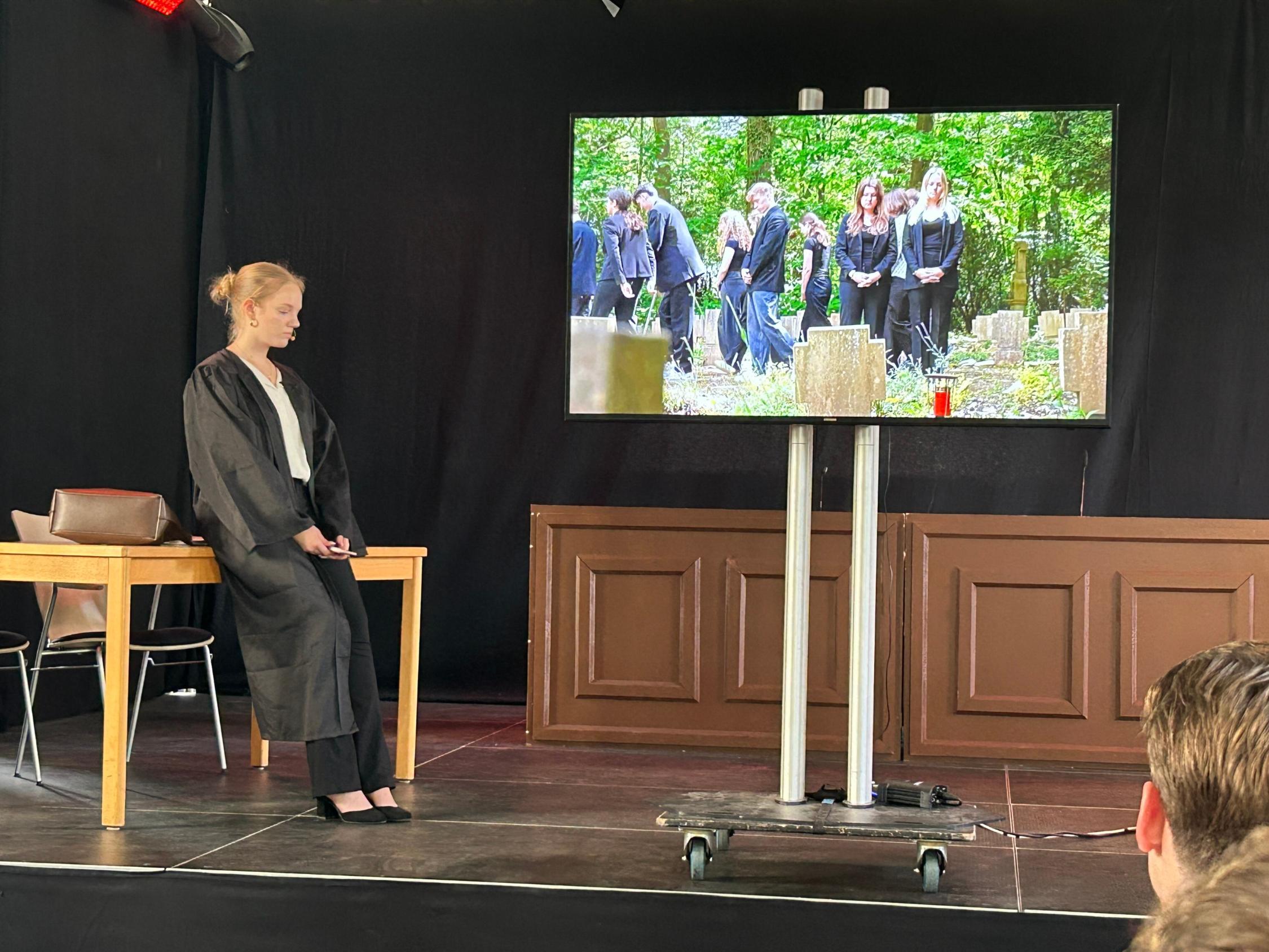 Monitor, darauf Beerdingunsszene, daneben die gespielte Rechtsanwältin