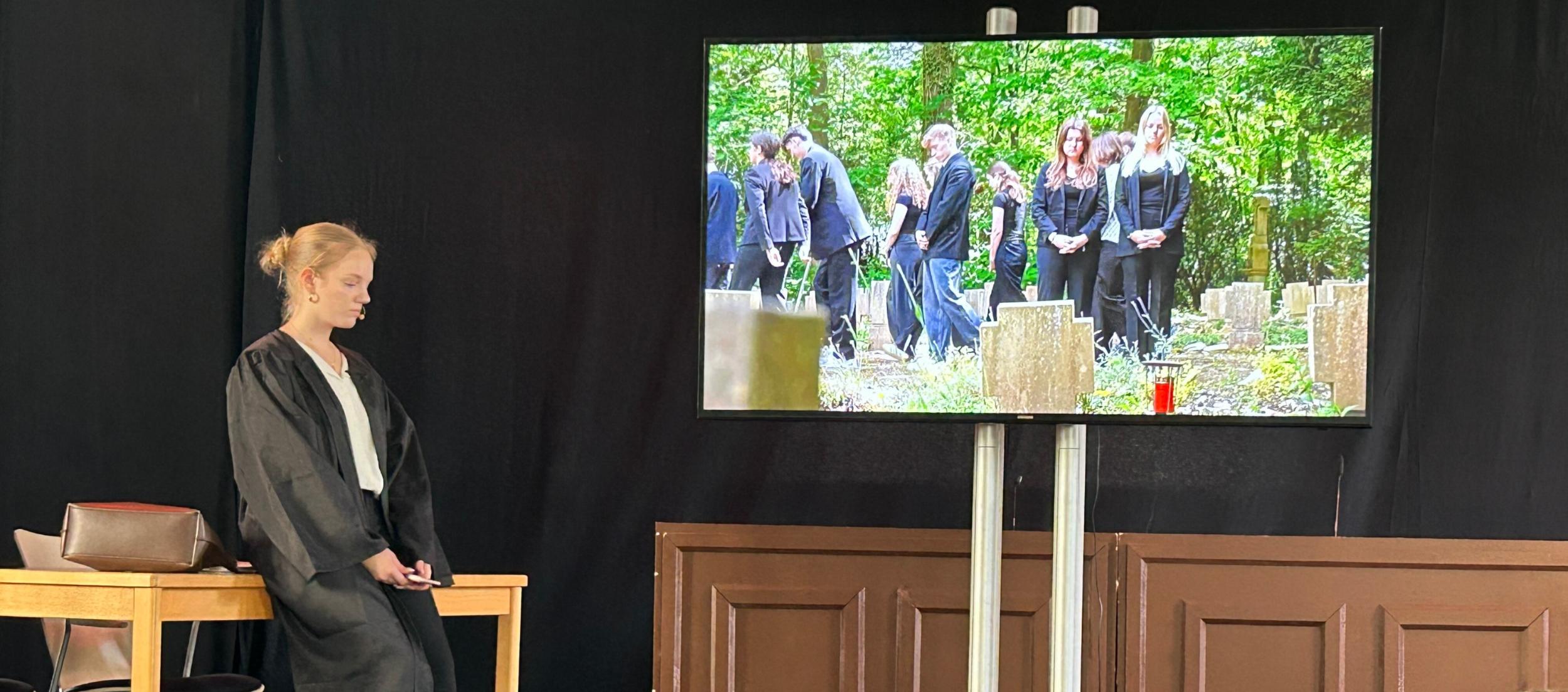 Monitor, darauf Beerdingunsszene, daneben die gespielte Rechtsanwältin