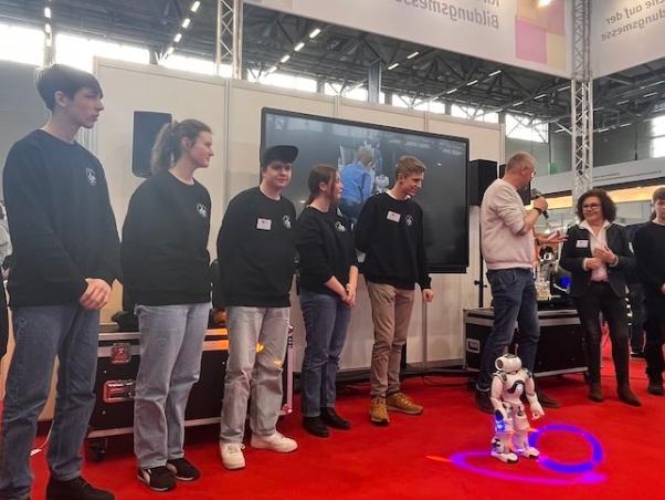 Schülerinnen und Schüler mit Technik-AG-Pullis sowie Lehrerin auf dem Messestand, davor ein Neo-Roboter