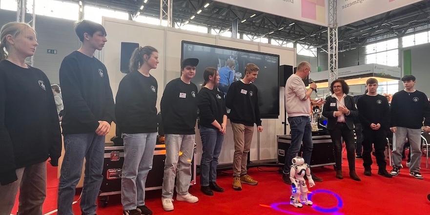 Schülerinnen und Schüler mit Technik-AG-Pullis sowie Lehrerin auf dem Messestand, davor ein Neo-Roboter