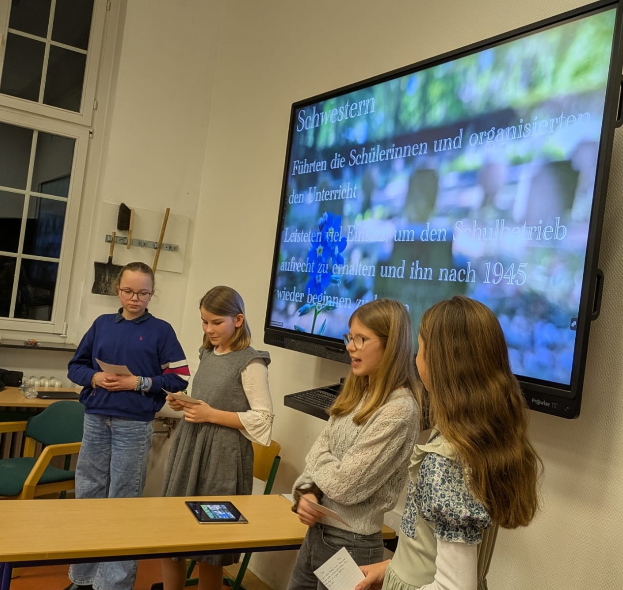 vier Mädchen vor einer digitalen Tafel, ein Mädchen spricht, auf der Tafel Infos über Schwestern