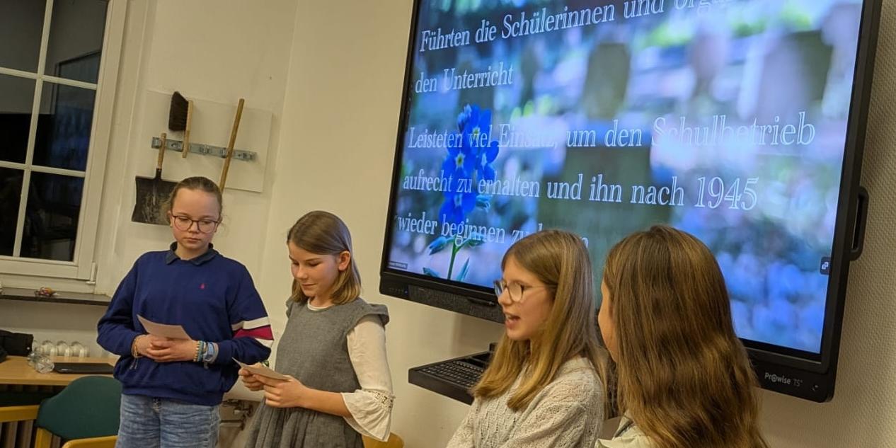 vier Mädchen vor einer digitalen Tafel, ein Mädchen spricht, auf der Tafel Infos über Schwestern