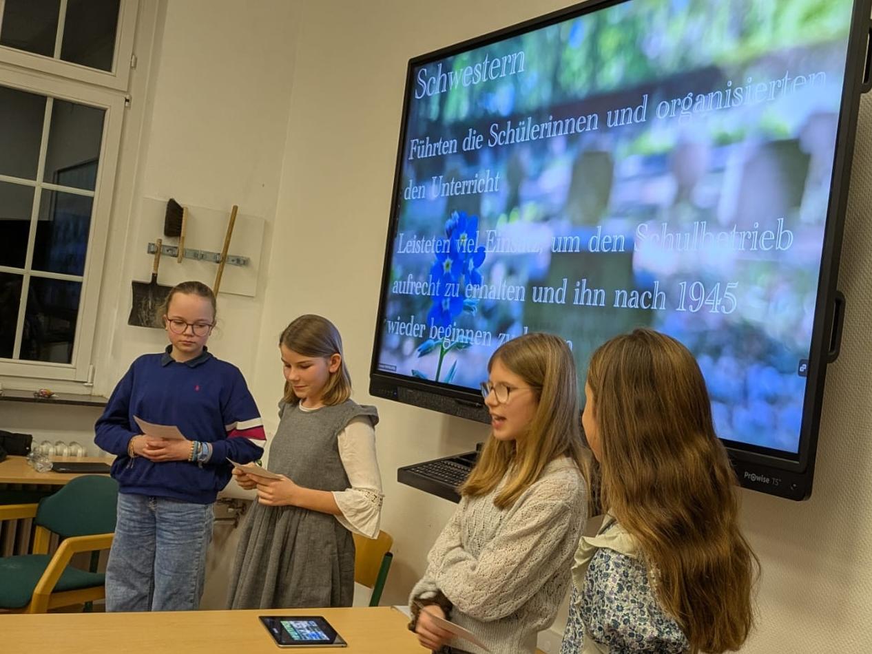 vier Mädchen vor einer digitalen Tafel, ein Mädchen spricht, auf der Tafel Infos über Schwestern