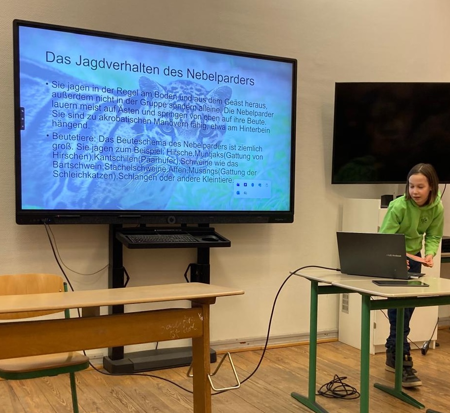 Mädchen am Laptop, daneben Präsentationswand: 'Das Jagdverhalten des Nebelparders'