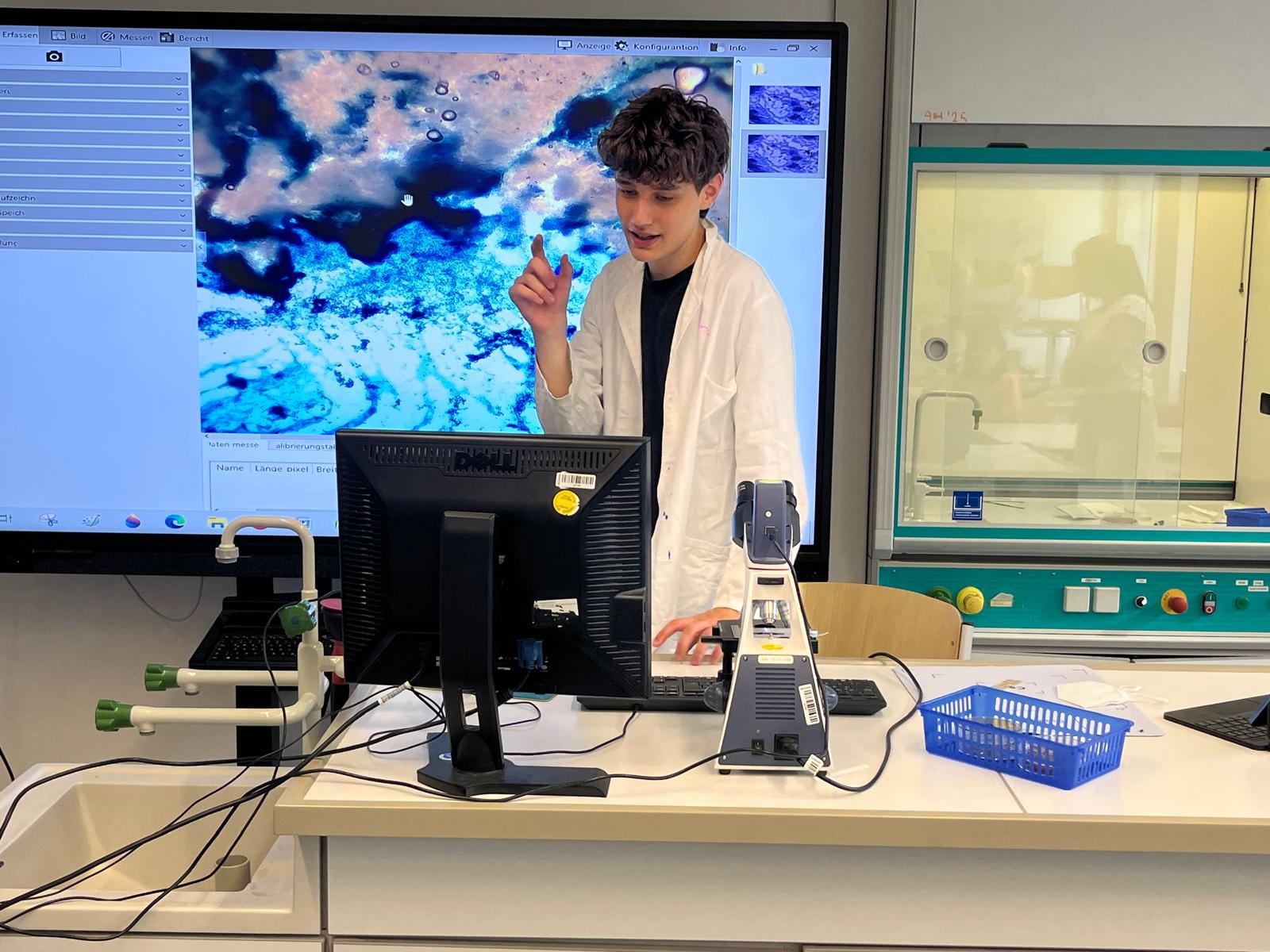 Schüler im Kittel erklärt ein Experiment, im Hintergrund Präsentation an einer digitalen Tafel