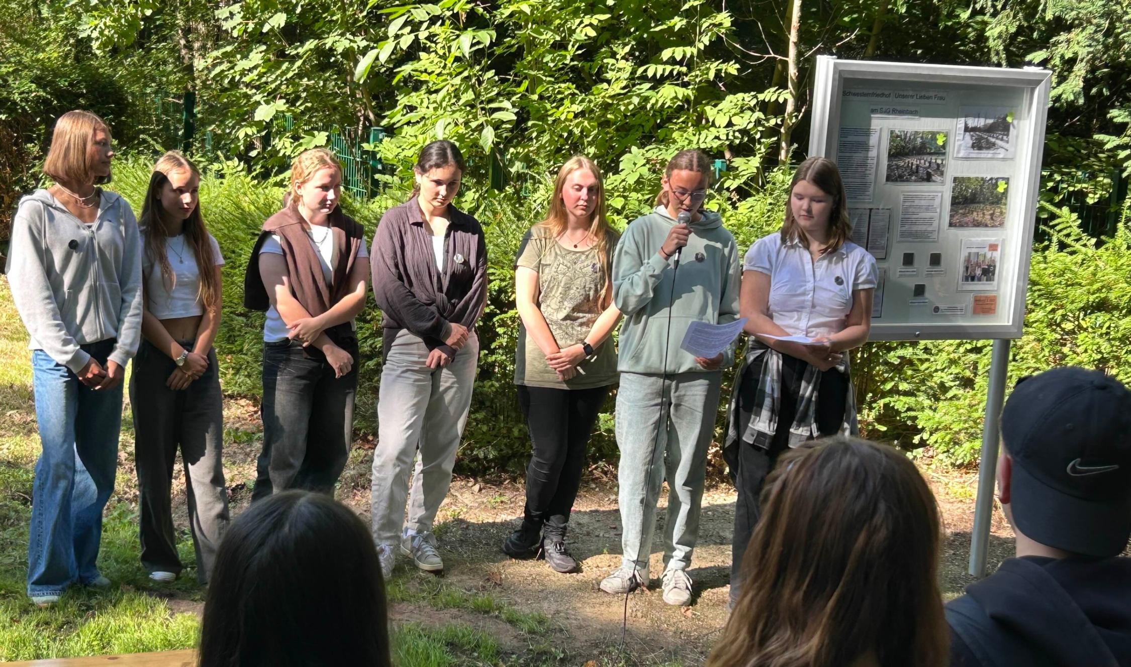 Schülerinnen stehen nebeneinander, eine hat ein Mikro in der Hand, rechts ein Schaukasten