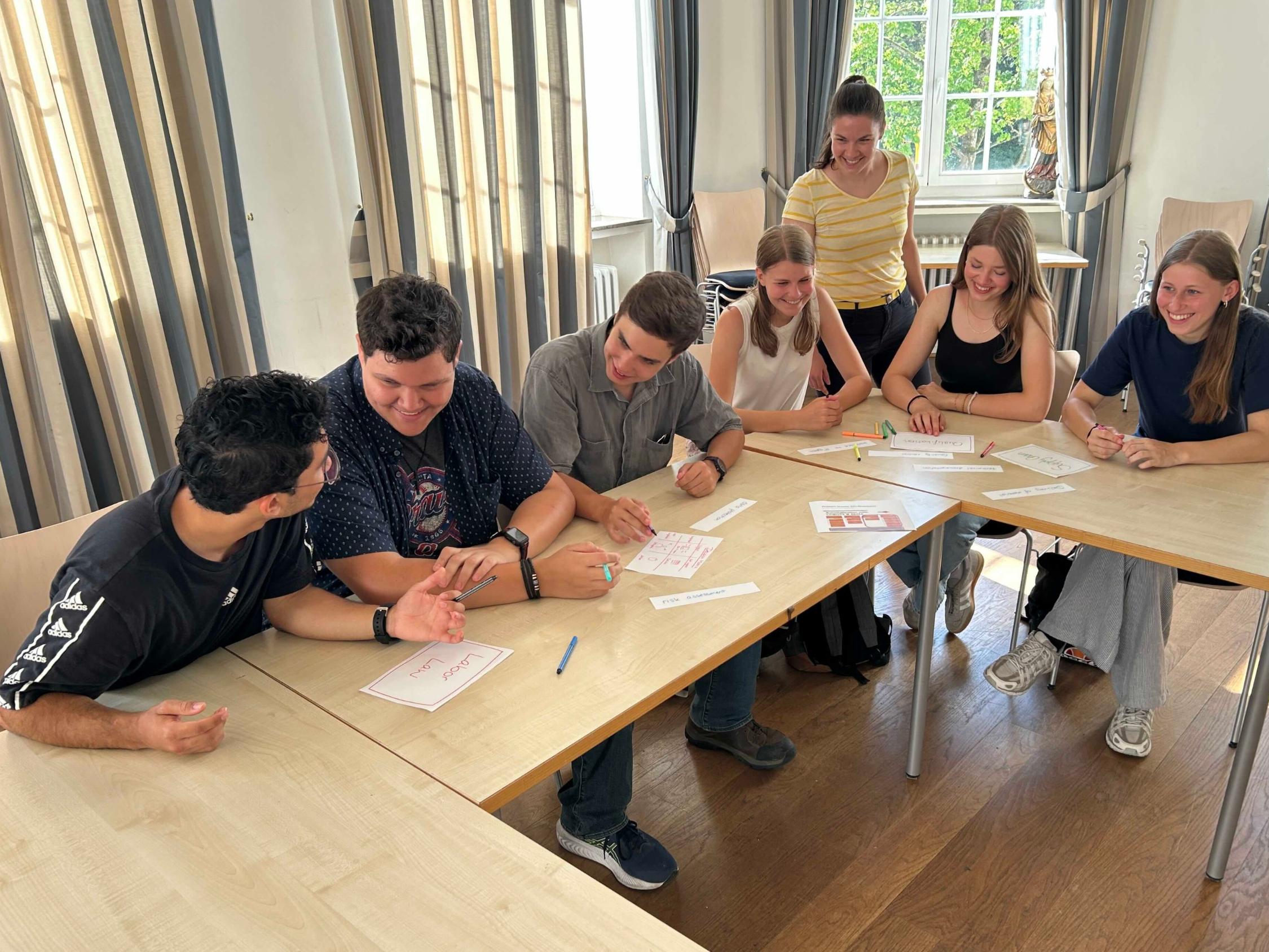 Schülerinnen und Schüler an Tischen im Gespräch, eine Lehrerin steht dabei