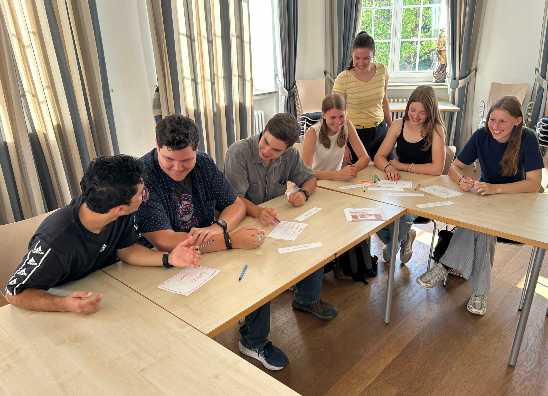 Schülerinnen und Schüler an Tischen im Gespräch, eine Lehrerin steht dabei