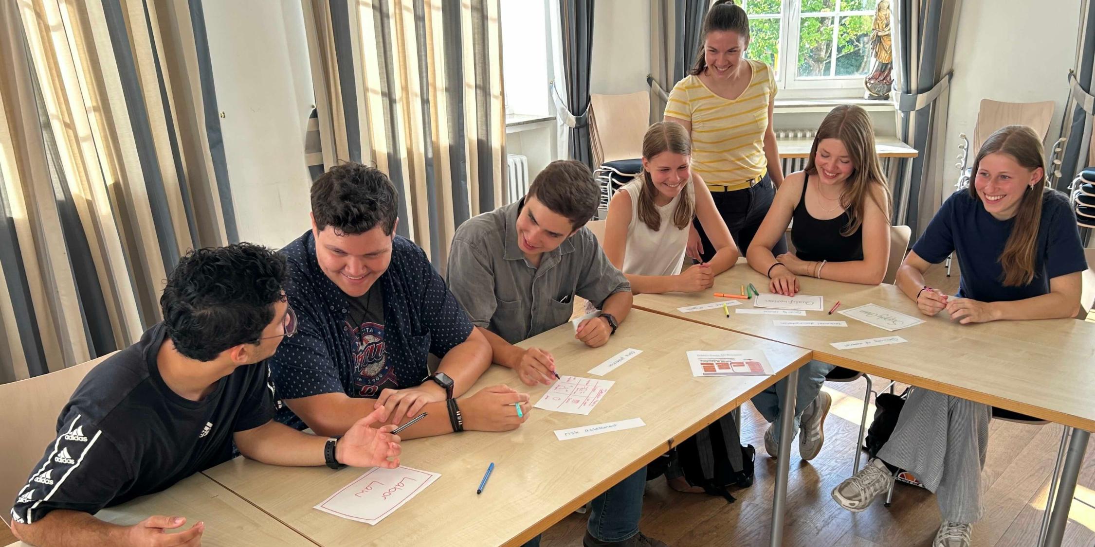 Schülerinnen und Schüler an Tischen im Gespräch, eine Lehrerin steht dabei