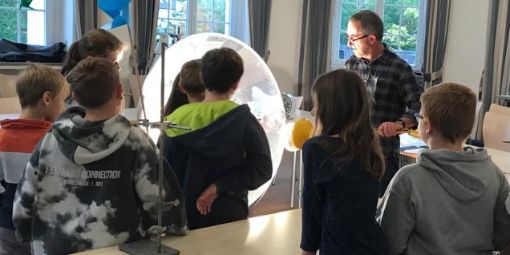 Gruppe aus Schüler:innen hört Dr. Gaßmann zu, der vor einer großen Metallschüssel das Experiment erklärt.