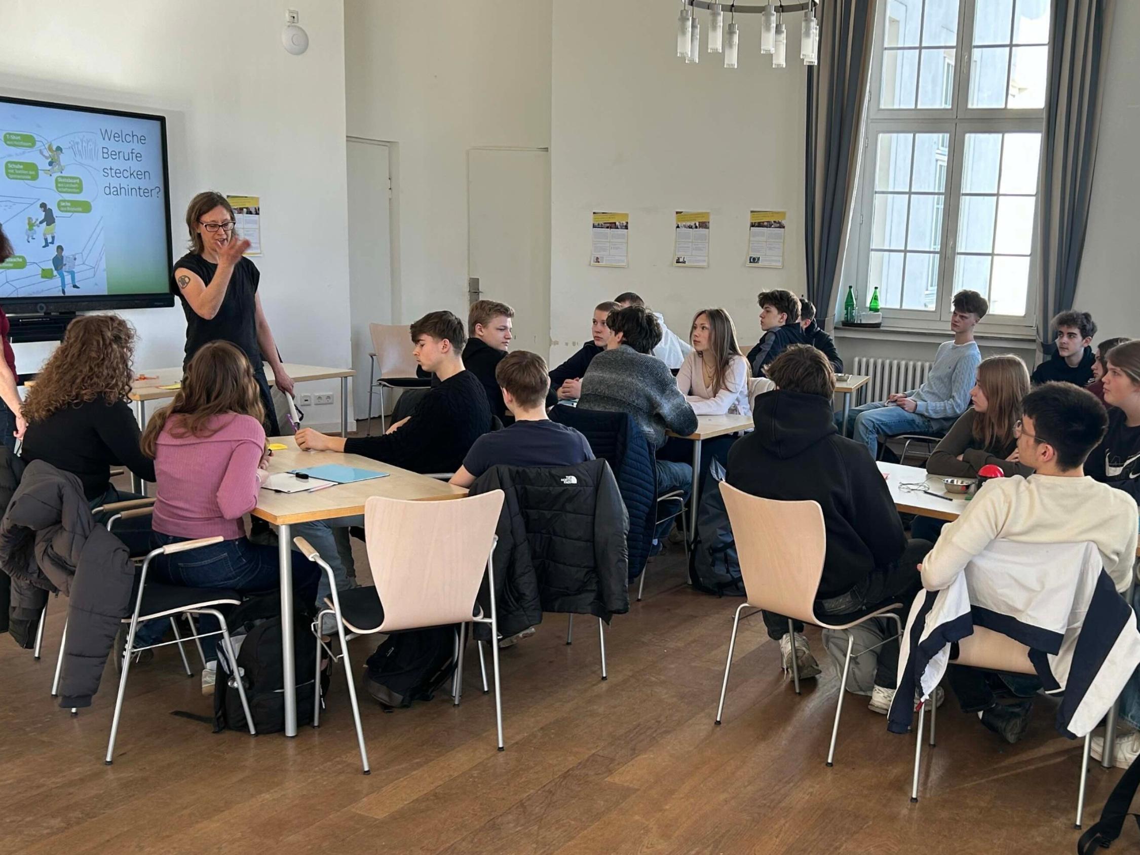 Schüler:innen sitzen an Gruppentischen, links ein Monitor, davor stehend zwei Frauen