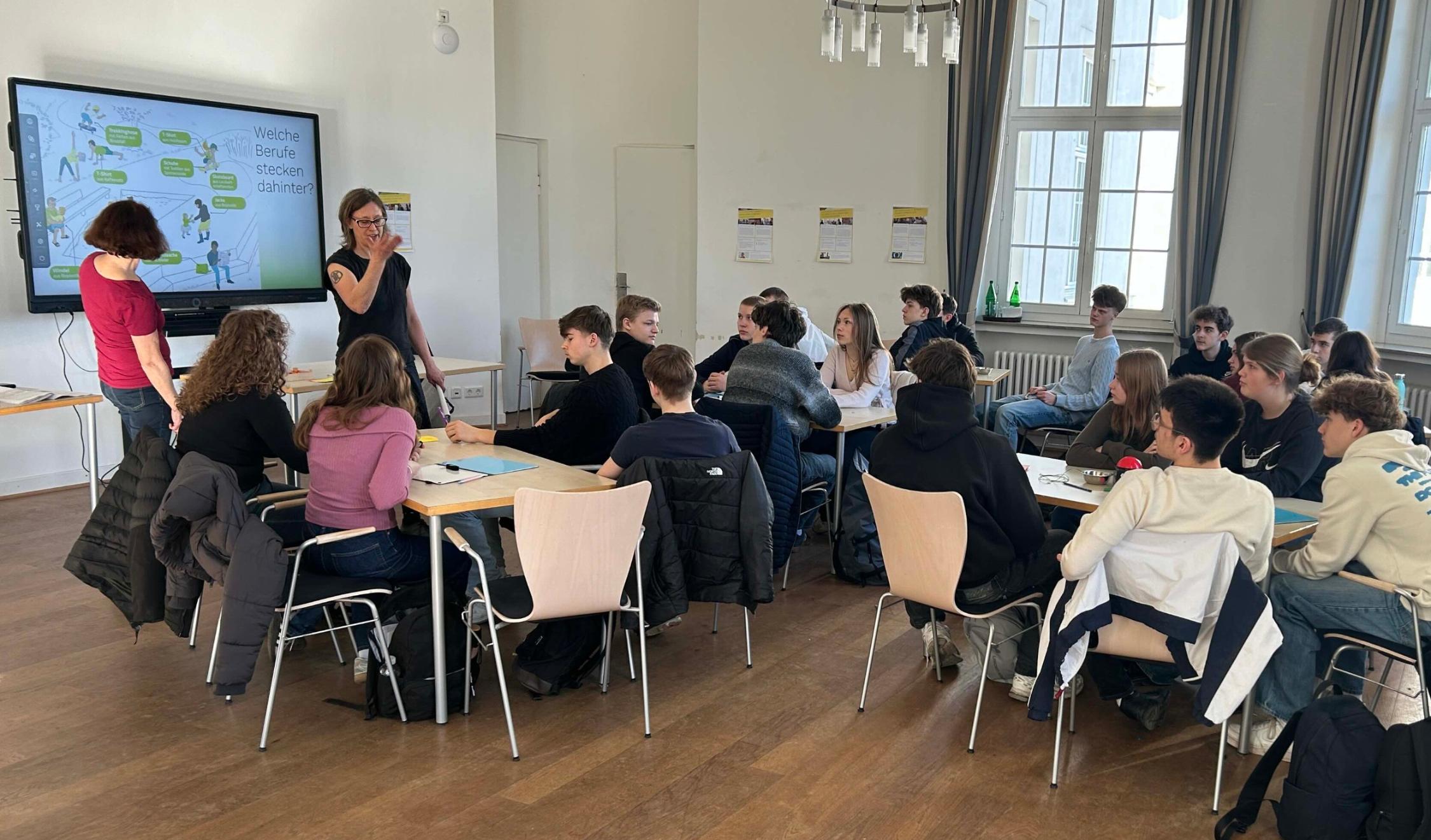 Schüler:innen sitzen an Gruppentischen, links ein Monitor, davor stehend zwei Frauen