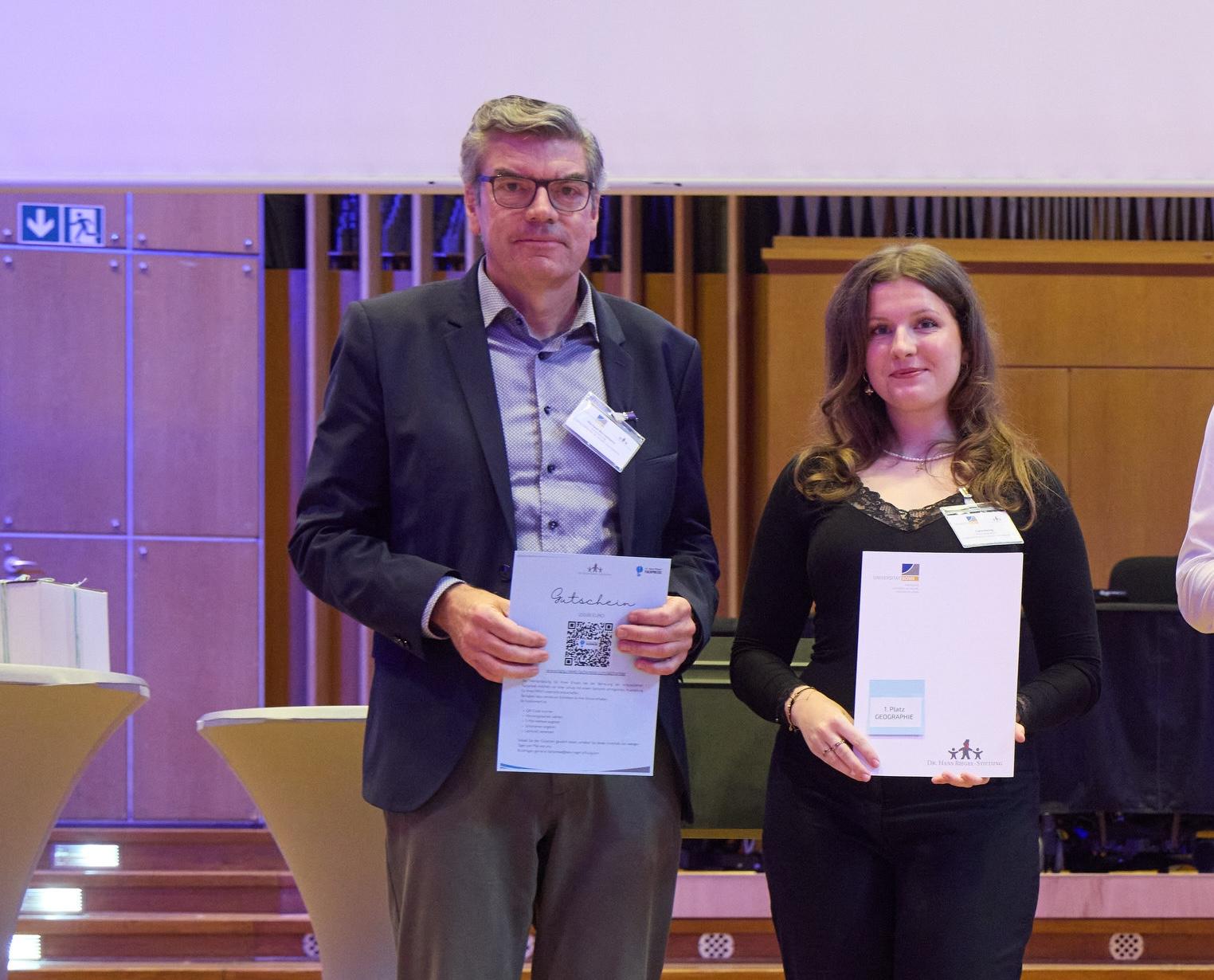 Mann (Schulleiter) und Schülerin mit Urkunden in der Hand