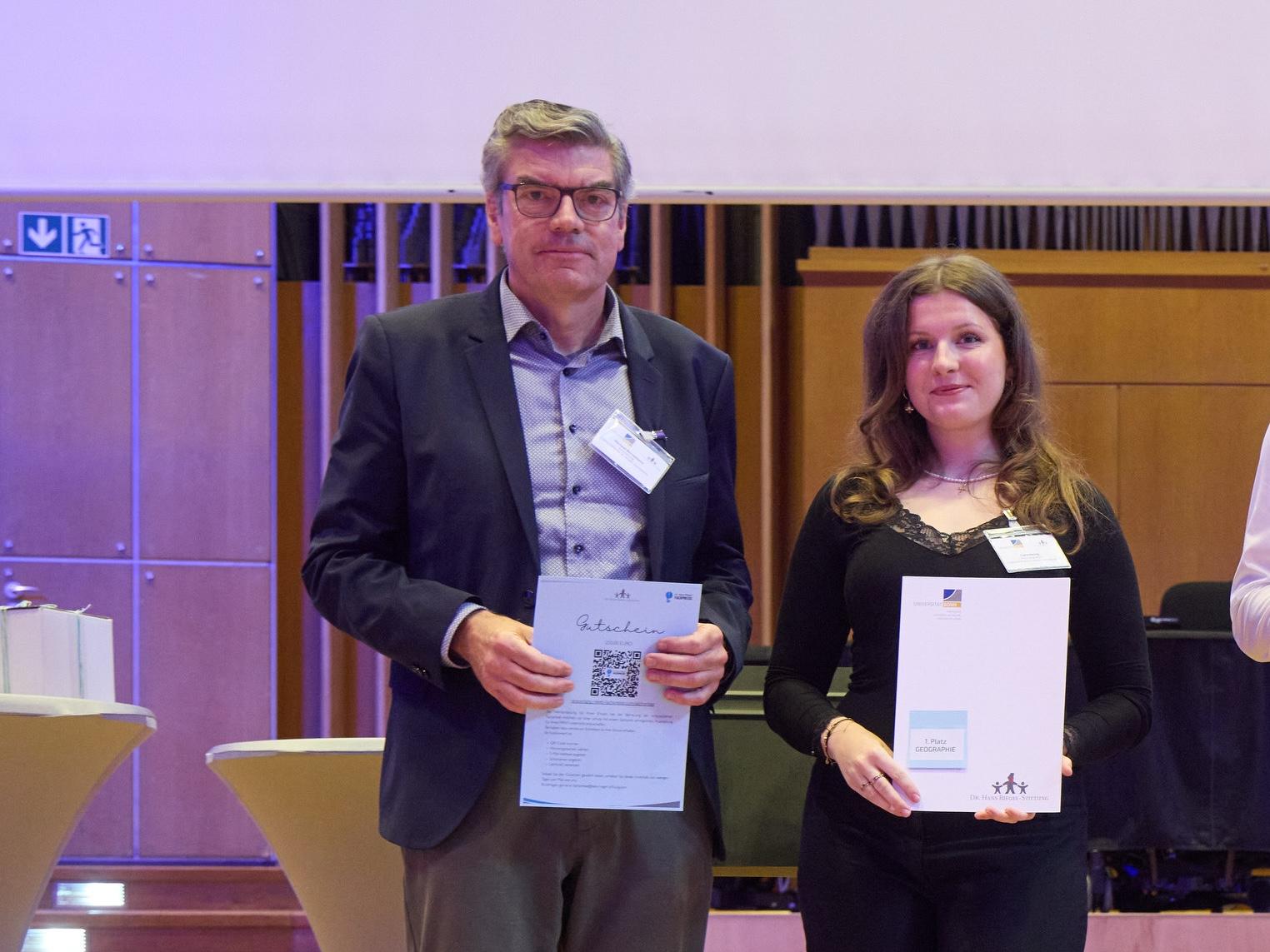 Mann (Schulleiter) und Schülerin mit Urkunden in der Hand