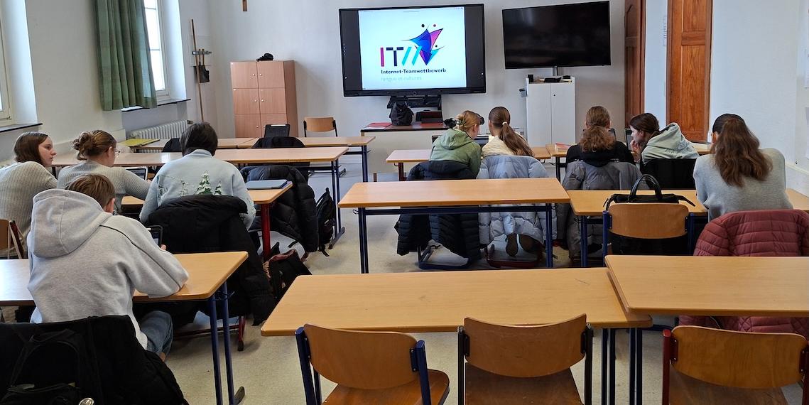 Schüler:innen von hinten in einem Klassenraum, digitales Board mit Schriftzug 'Internet Team Wettbewerb'