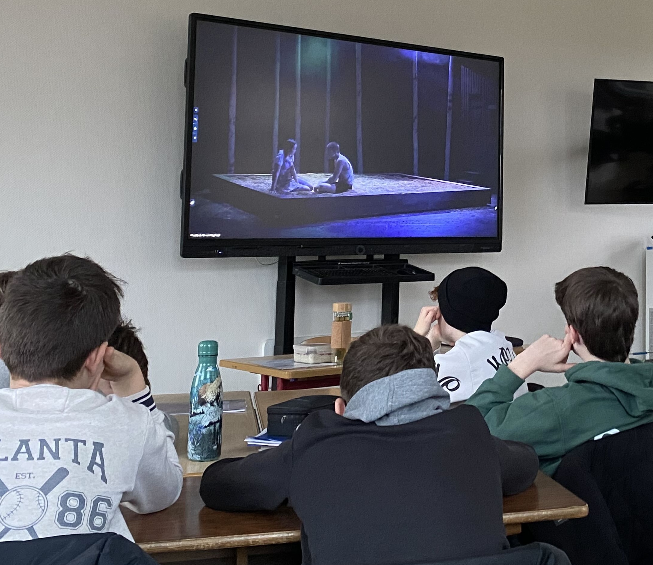 Bildschirm: zwei Jugendliche sitzen auf dem Boden. Vor dem Bildschirm Jungen sitzend im Klassenraum