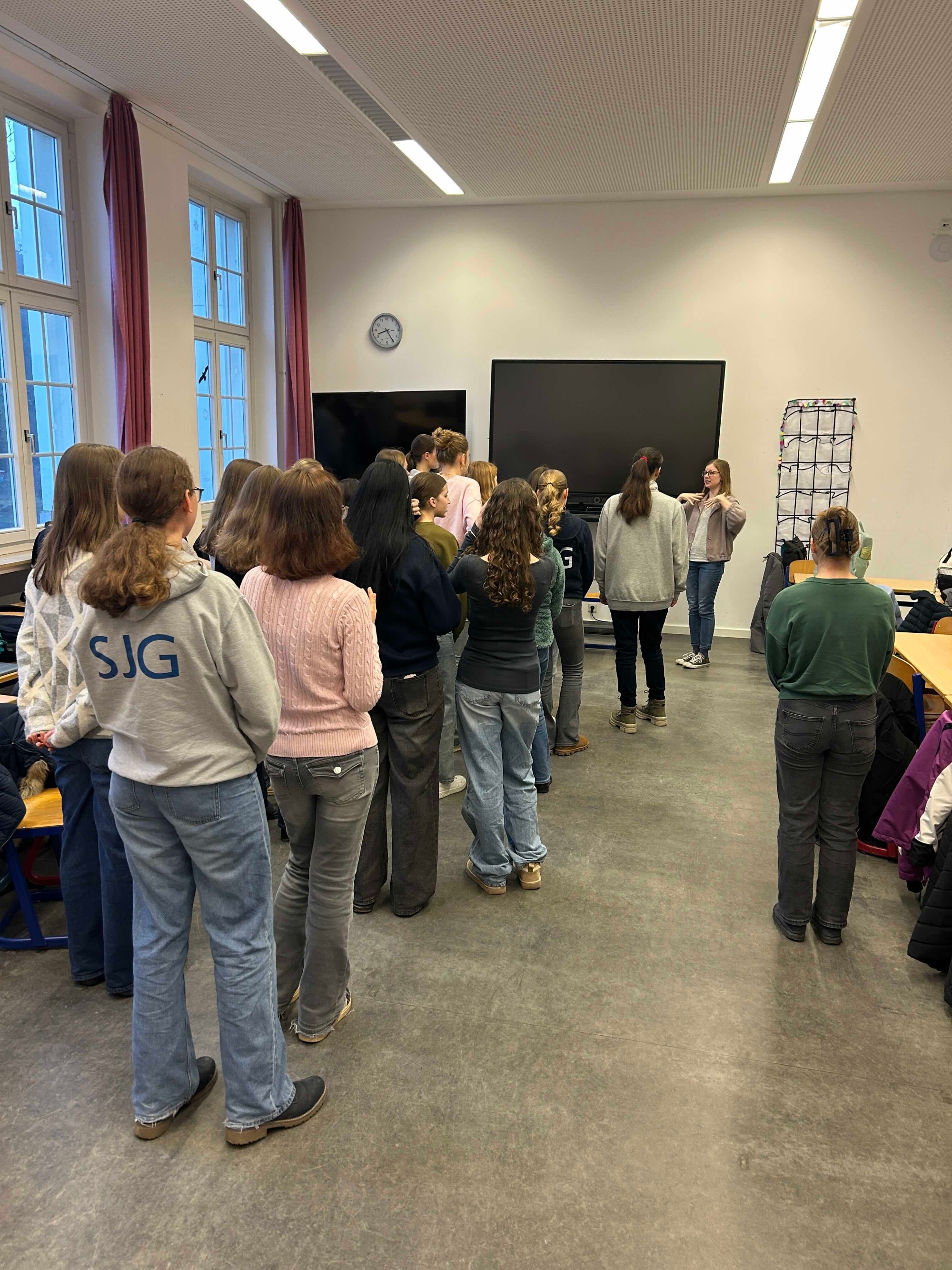 Mädchen stehen in der Klasse und blicken zu einer Frau, die eine Bewegung anzeigt