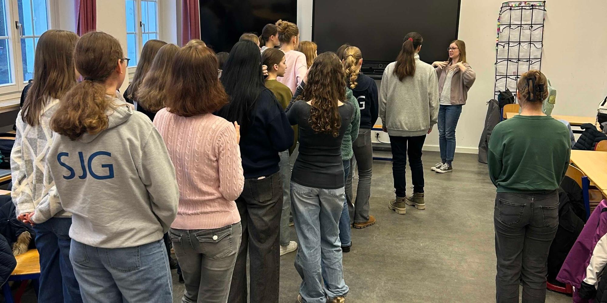 Mädchen stehen in der Klasse und blicken zu einer Frau, die eine Bewegung anzeigt