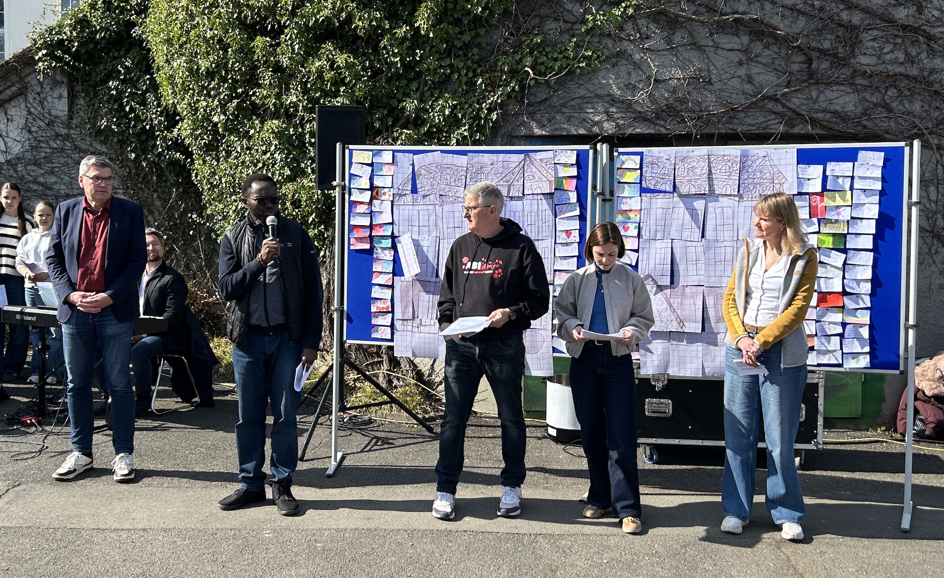 fünf Erwachsene stehen vor einer Stellwand, ein Priester spricht