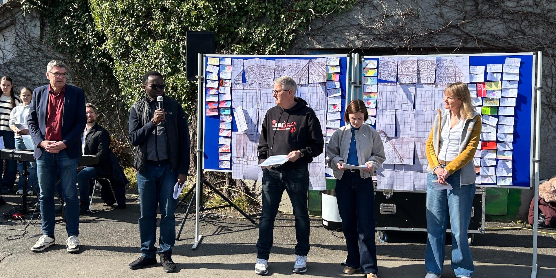 fünf Erwachsene stehen vor einer Stellwand, ein Priester spricht