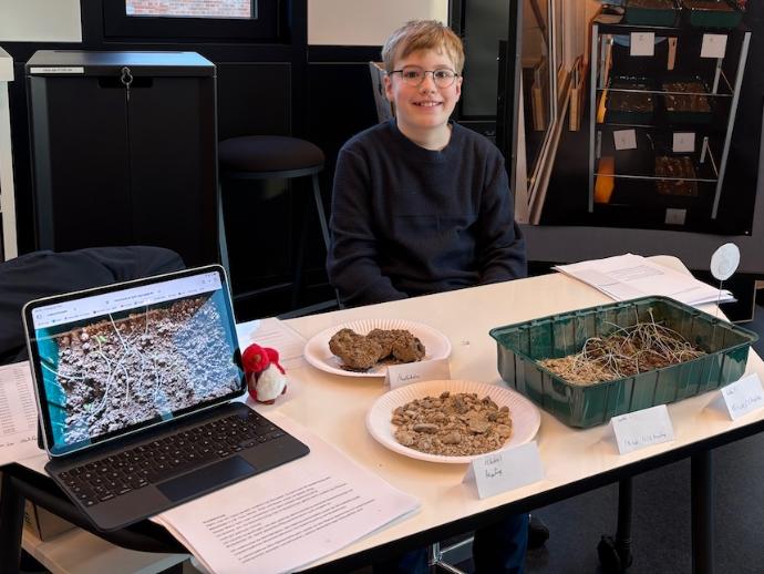 Junge sitzt hinter einem Tisch, auf dem Tisch ein aufgeklapptes Notebook, Betonkrümel, Pflanzenschale