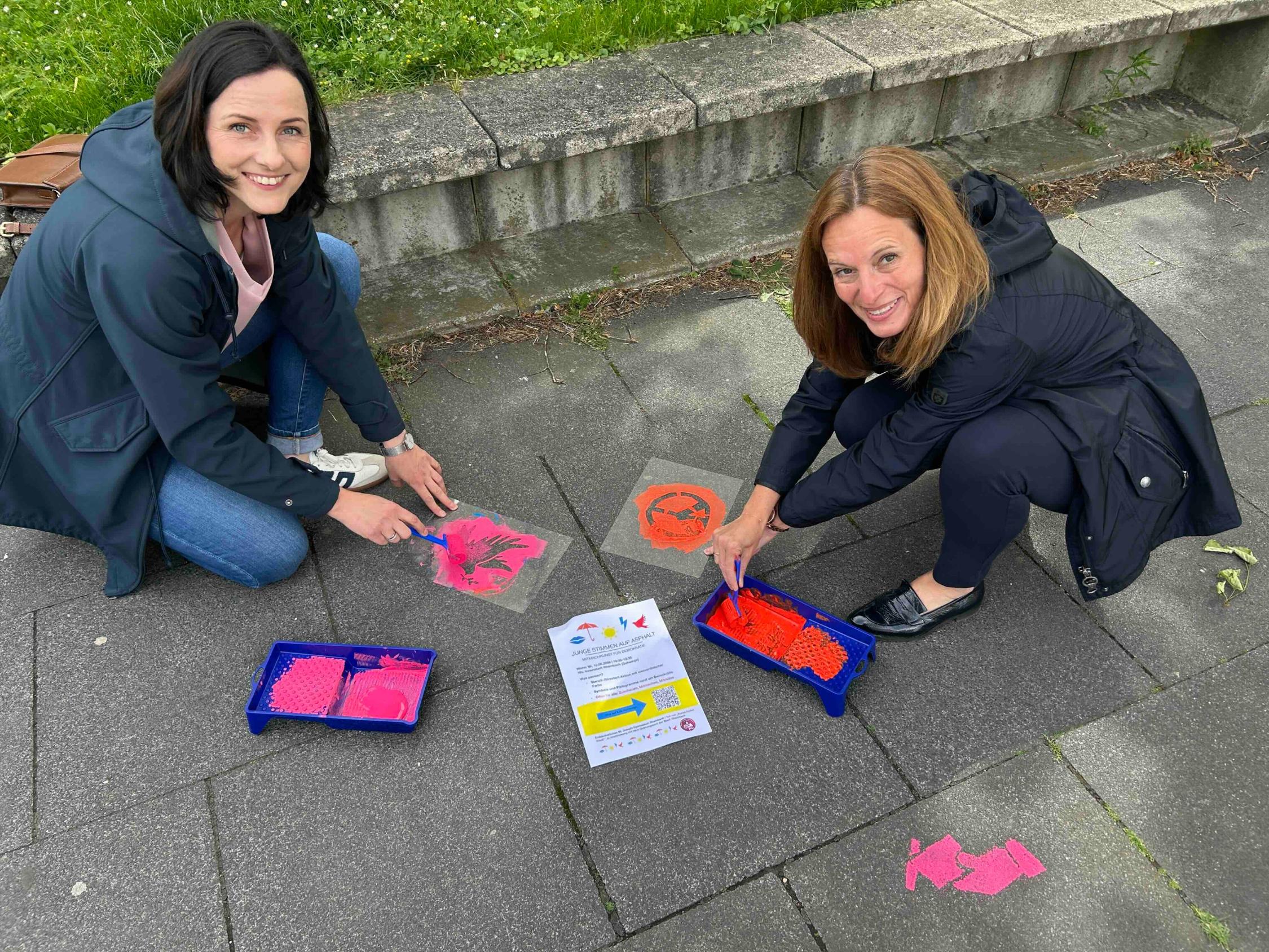 zwei Frauen arbeiten mit Farbe und Schablonen auf dem Gehweg