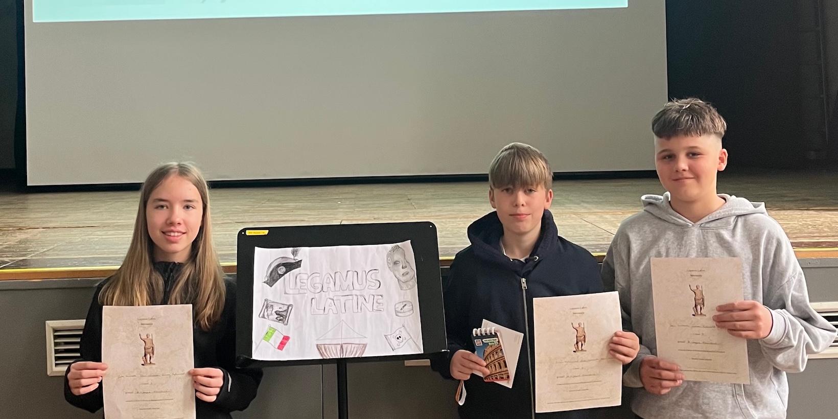 zwei Schüler, eine Schülerin mit Urkunden in der Hand, über ihnen projiziert: Legamus Latina