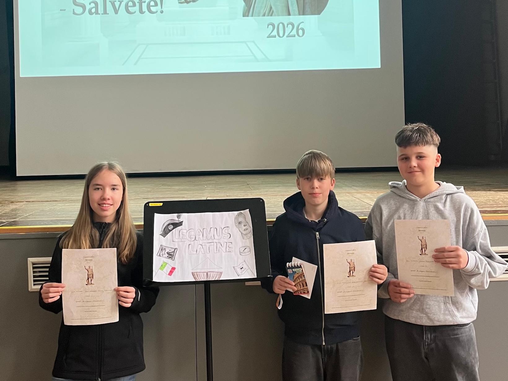 zwei Schüler, eine Schülerin mit Urkunden in der Hand, über ihnen projiziert: Legamus Latina