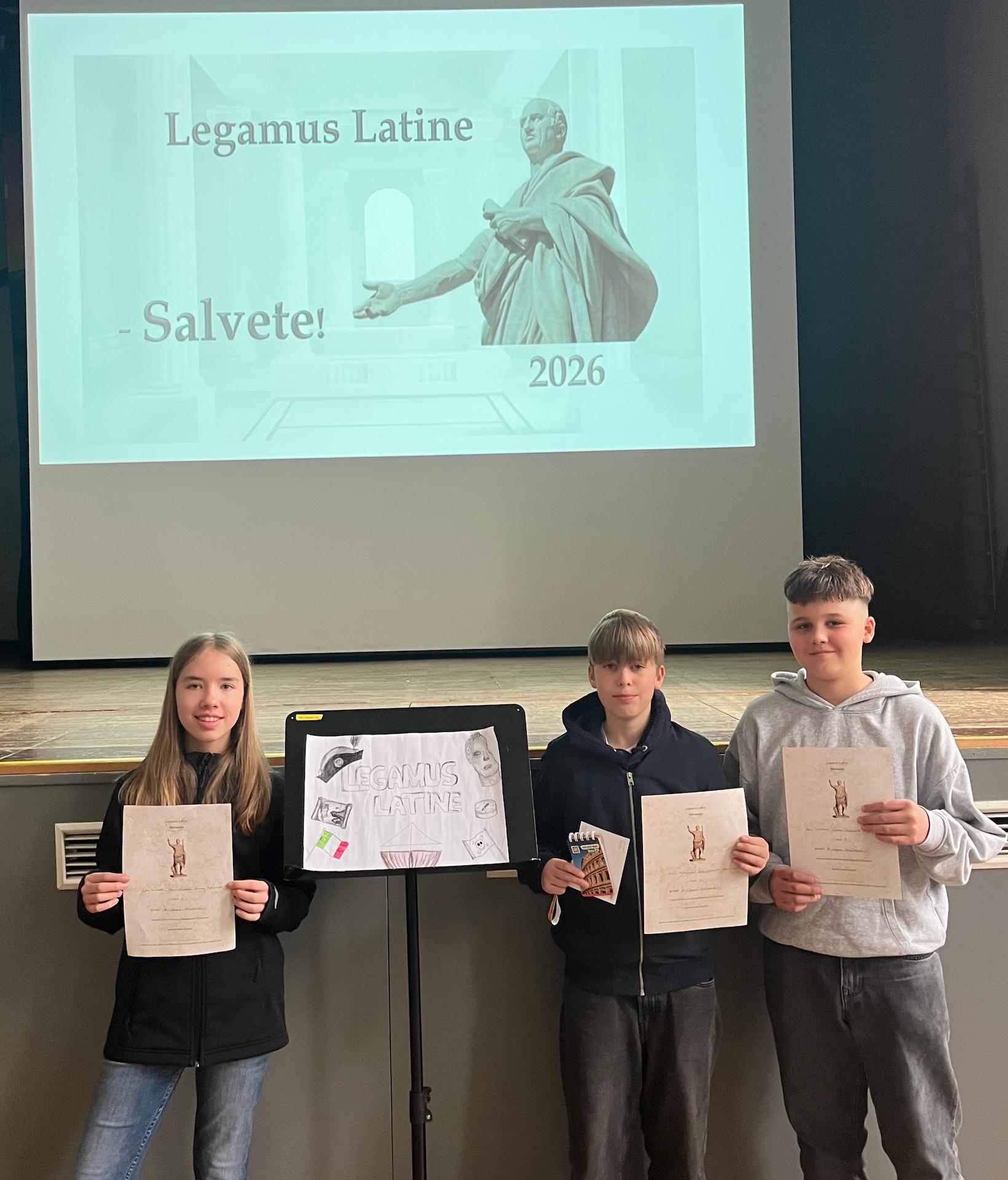 zwei Schüler, eine Schülerin mit Urkunden in der Hand, über ihnen projiziert: Legamus Latina