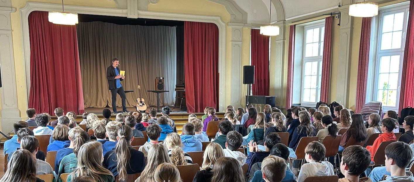 Schülerinnen und Schüler von hinten auf Stühlen mit Blick auf die Bühne, dort steht ein Mann mit einem Buch in der Hand