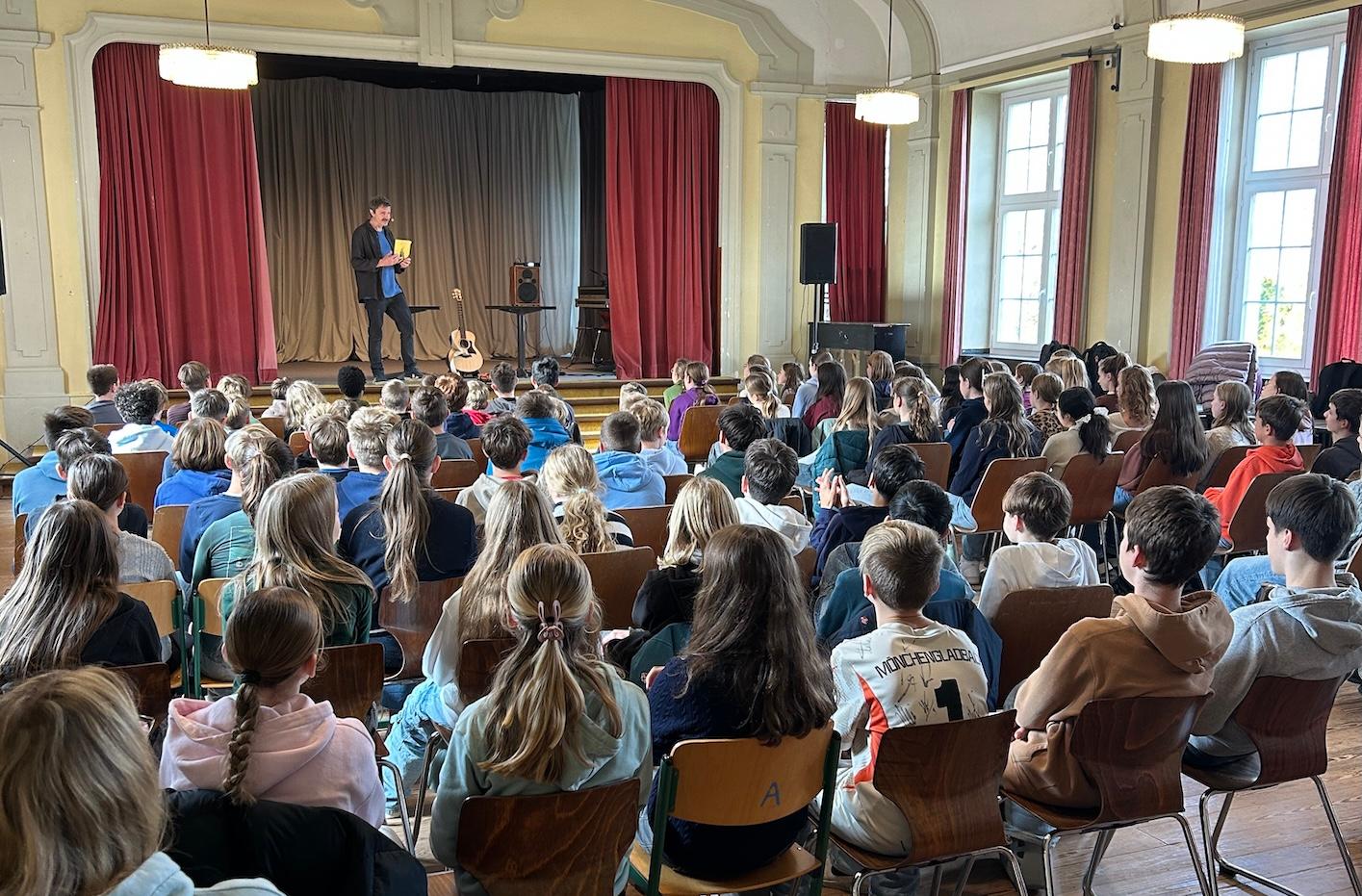 Schülerinnen und Schüler von hinten auf Stühlen mit Blick auf die Bühne, dort steht ein Mann mit einem Buch in der Hand