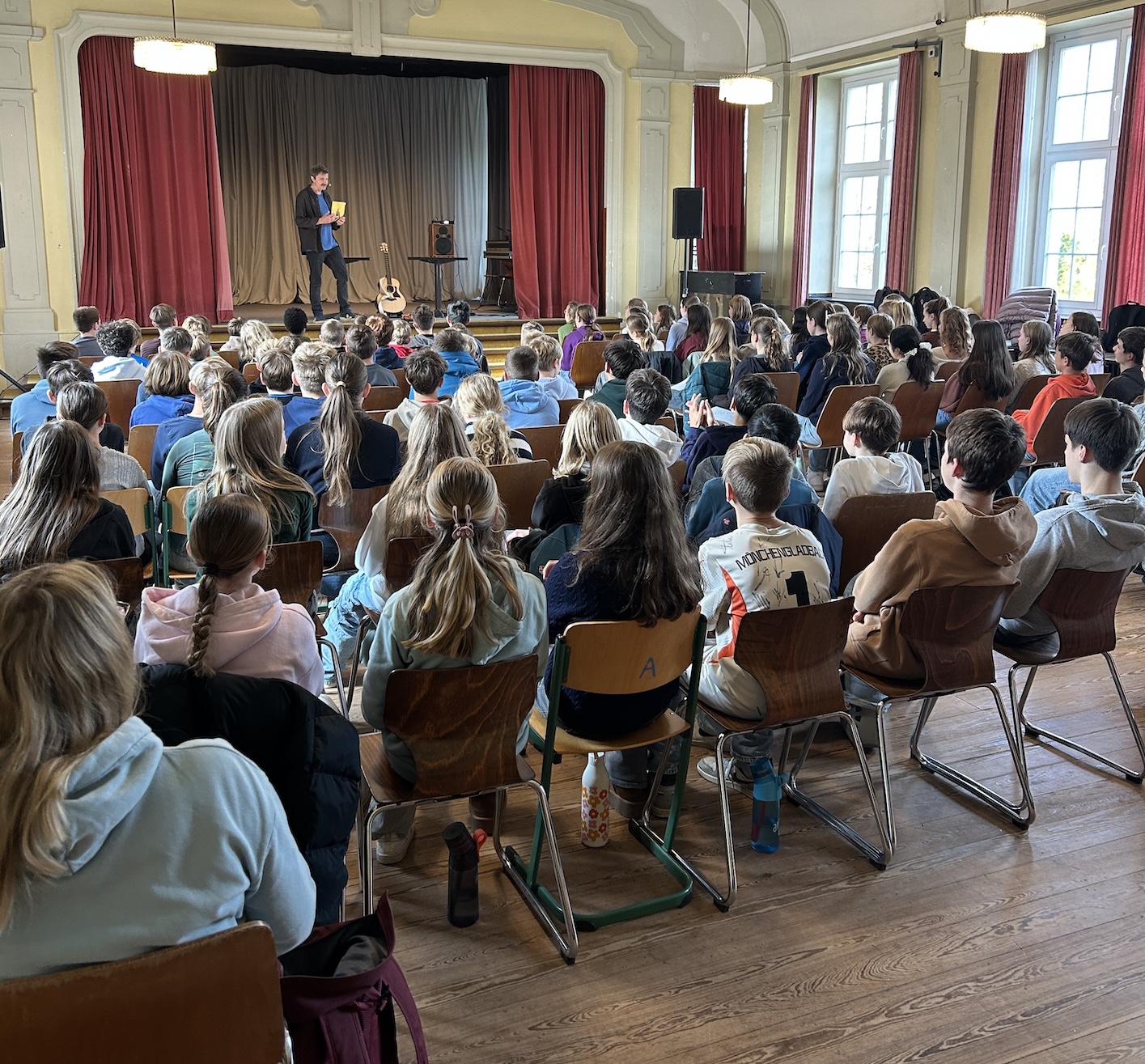 Schülerinnen und Schüler von hinten auf Stühlen mit Blick auf die Bühne, dort steht ein Mann mit einem Buch in der Hand
