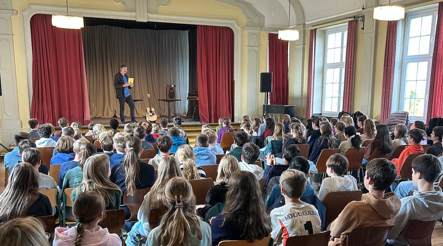 Schülerinnen und Schüler von hinten auf Stühlen mit Blick auf die Bühne, dort steht ein Mann mit einem Buch in der Hand