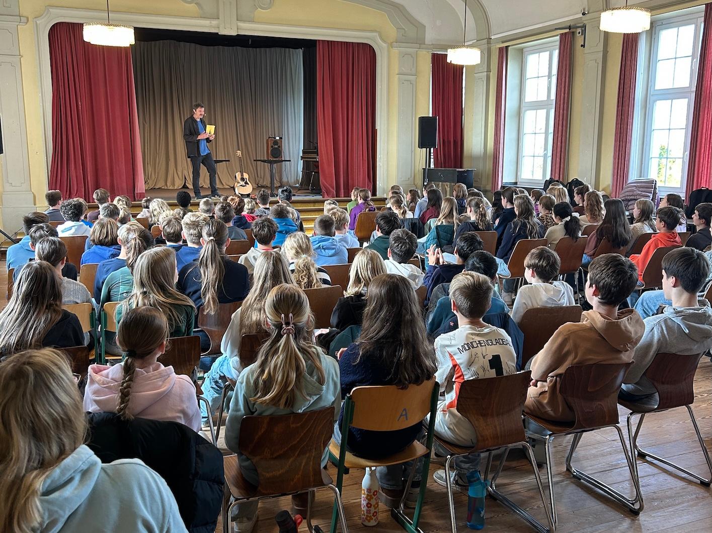 Schülerinnen und Schüler von hinten auf Stühlen mit Blick auf die Bühne, dort steht ein Mann mit einem Buch in der Hand