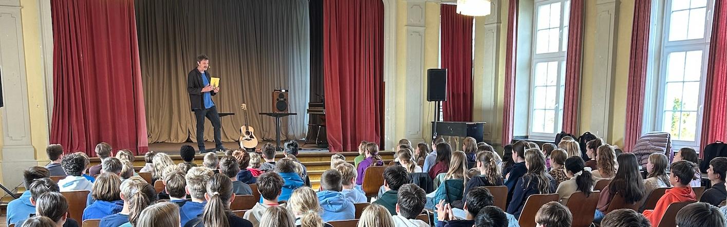 Schülerinnen und Schüler von hinten auf Stühlen mit Blick auf die Bühne, dort steht ein Mann mit einem Buch in der Hand