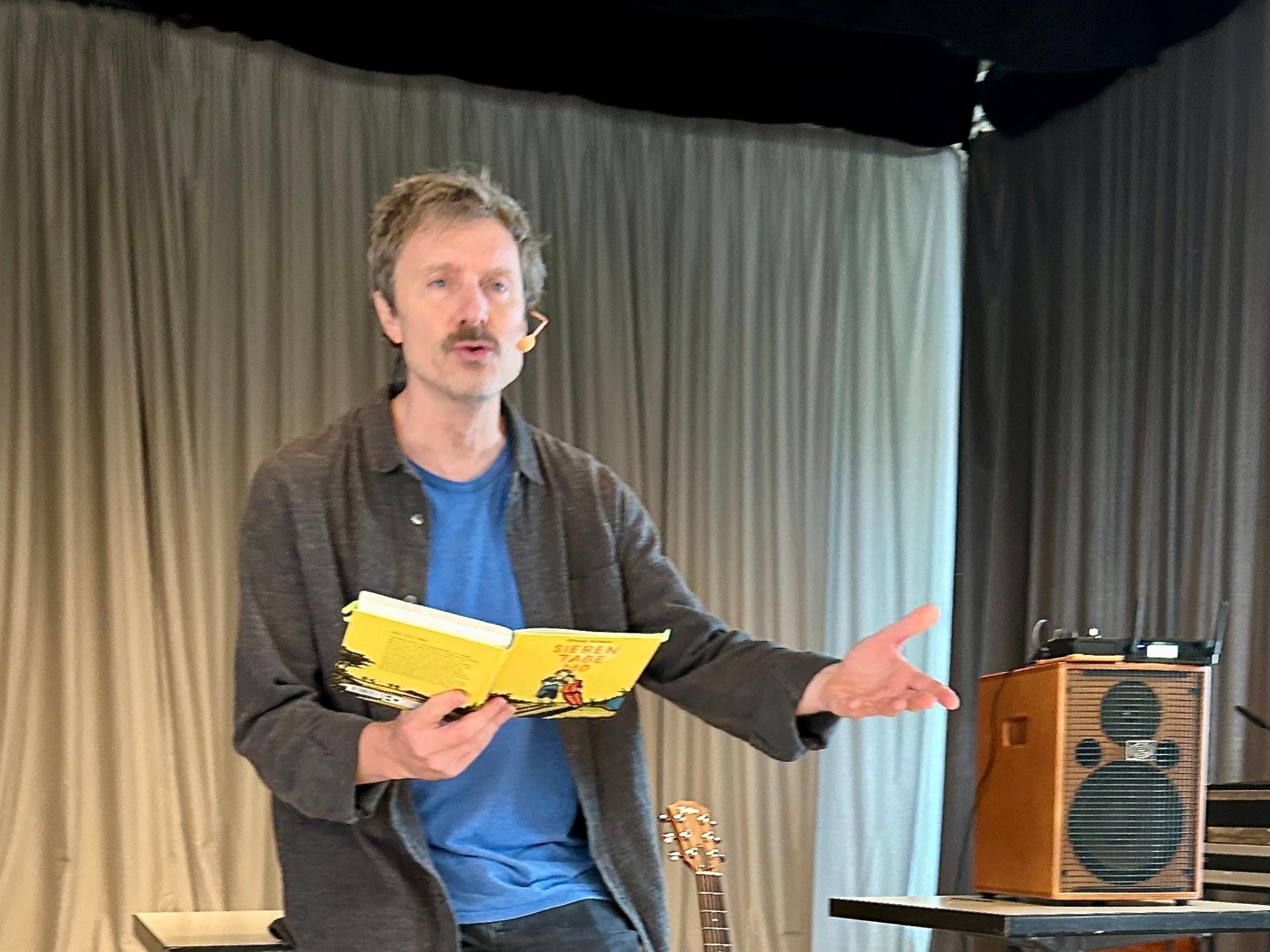 Man lehnt an einem Tisch, liest aus einem Buch, Hand ausgestreckt, neben ihm eine Gitarre und ein Verstärker