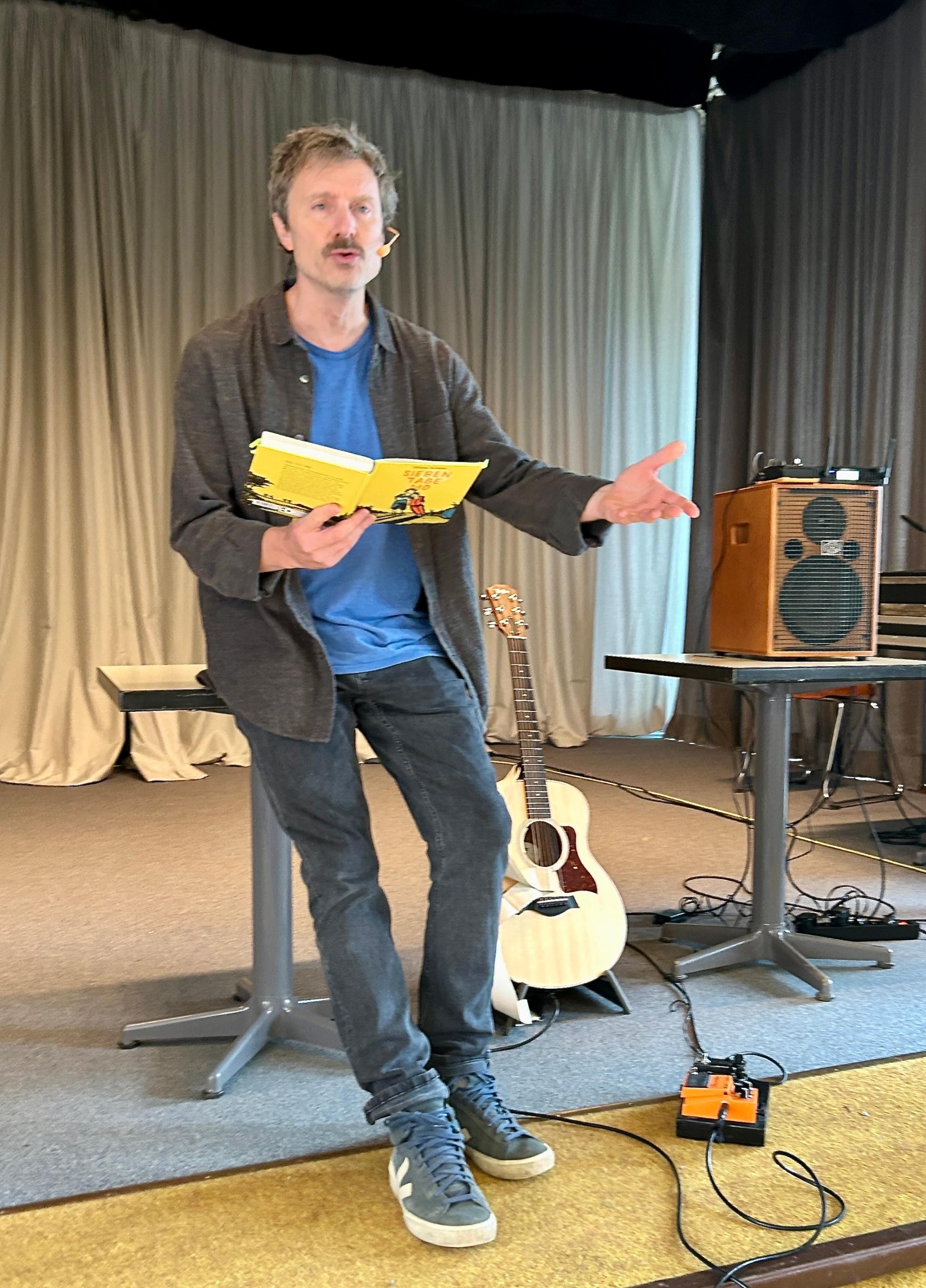 Man lehnt an einem Tisch, liest aus einem Buch, Hand ausgestreckt, neben ihm eine Gitarre und ein Verstärker