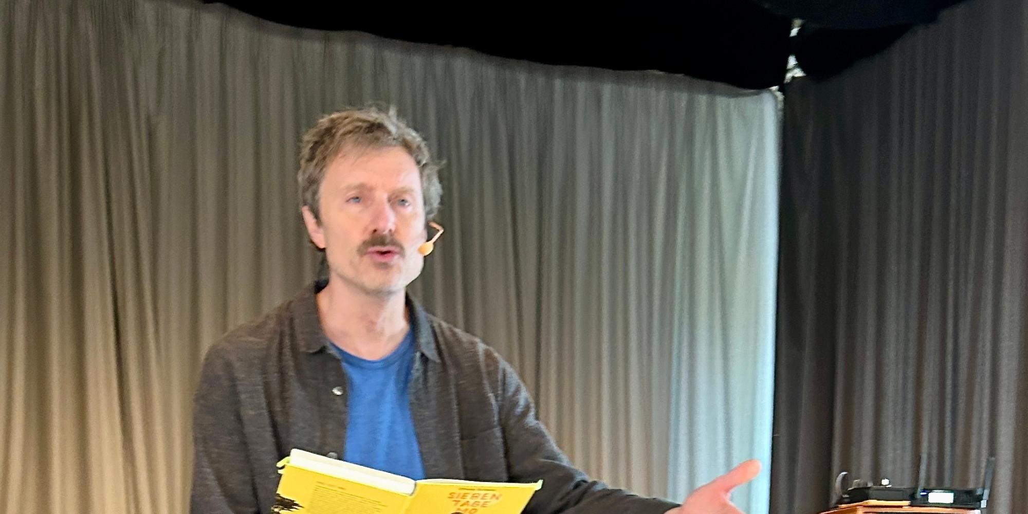 Man lehnt an einem Tisch, liest aus einem Buch, Hand ausgestreckt, neben ihm eine Gitarre und ein Verstärker